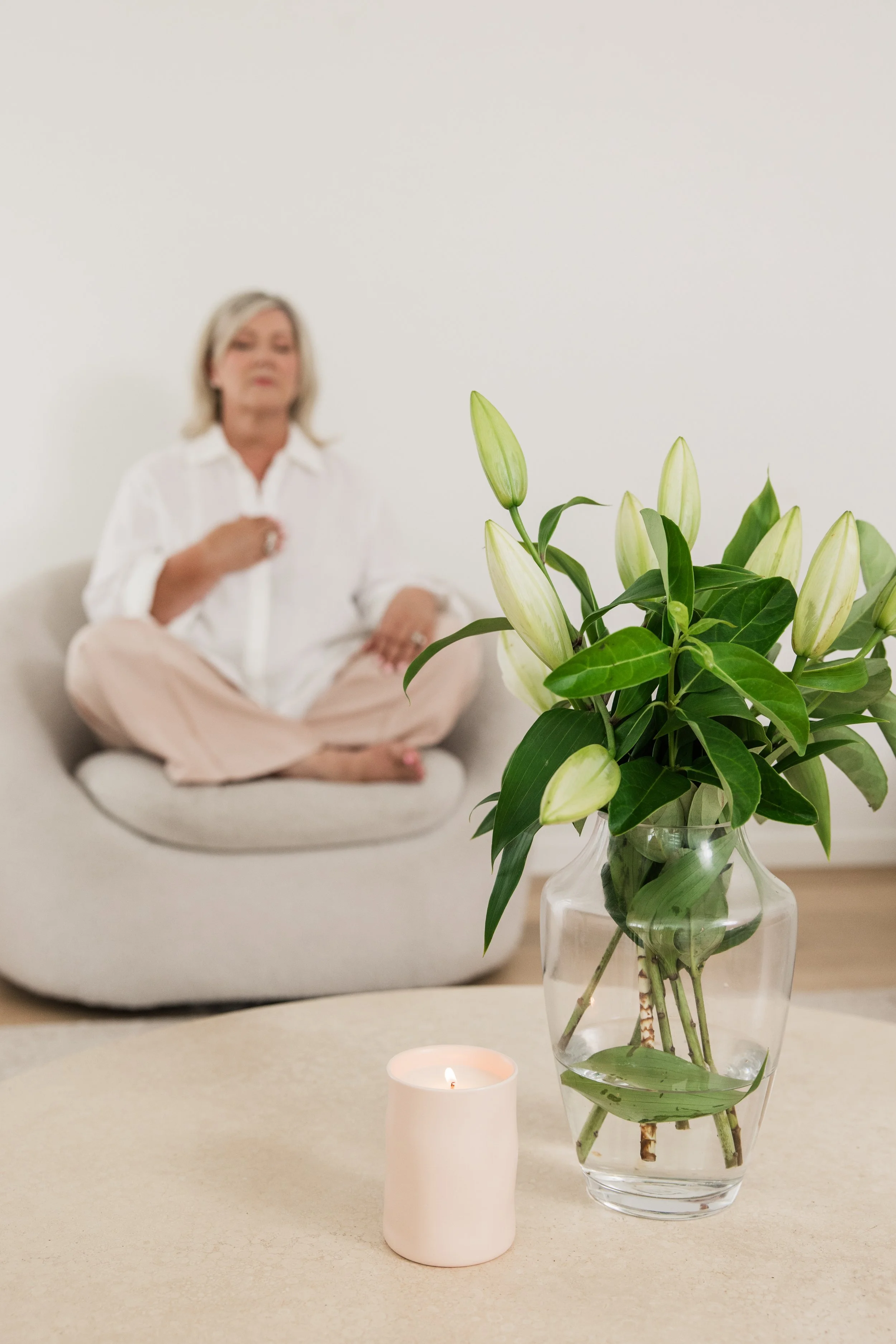 A white vase with white lilies on a table, a lit candle, with a woman sitting on a sofa in the background.
