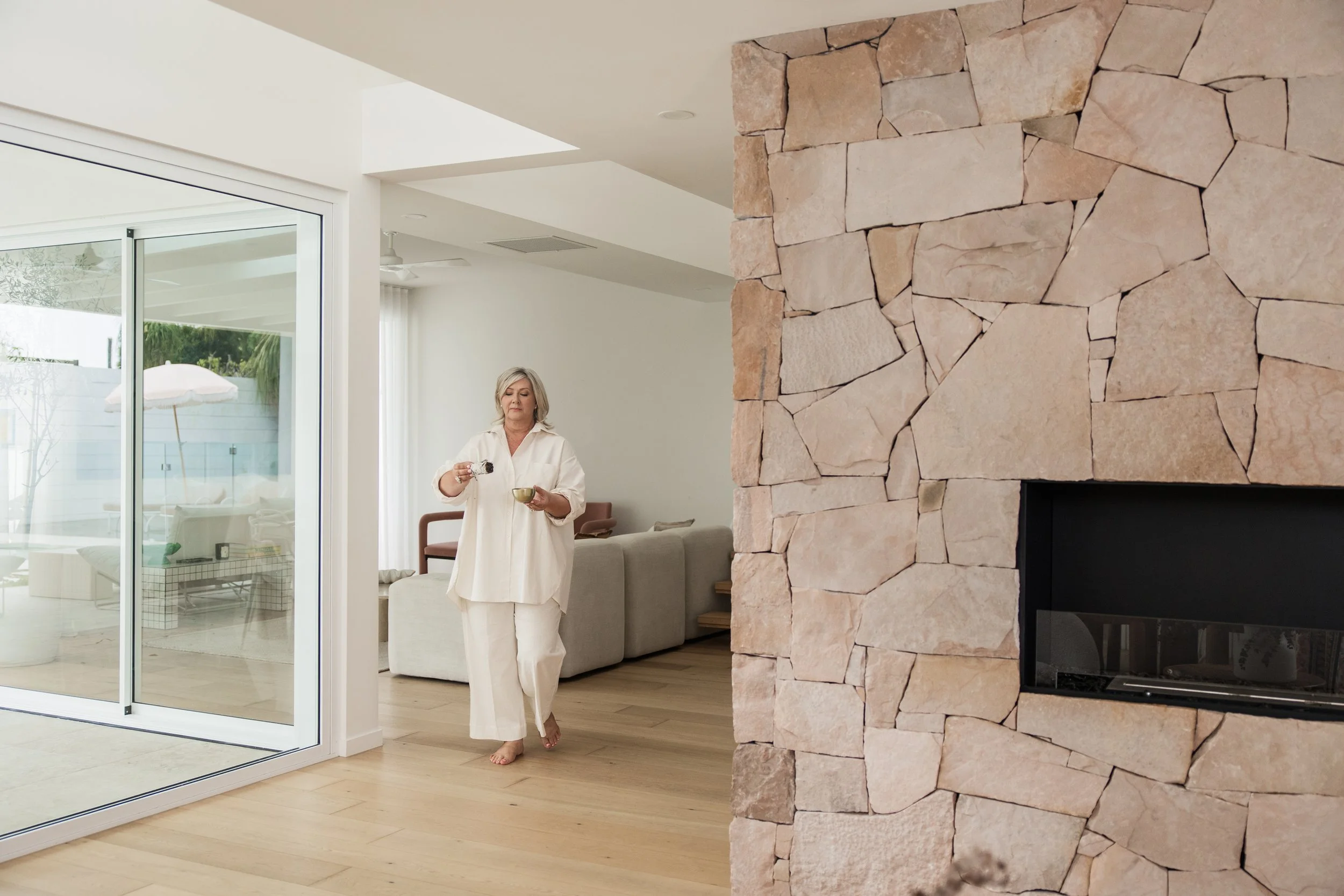 A woman with short blond hair, dressed in white pajamas, walking barefoot in a modern living room, holding a small bowl in one hand and a spoon in the other. The room features a stone fireplace on the right, a large glass sliding door on the left, and a neutral-colored sofa in the background.