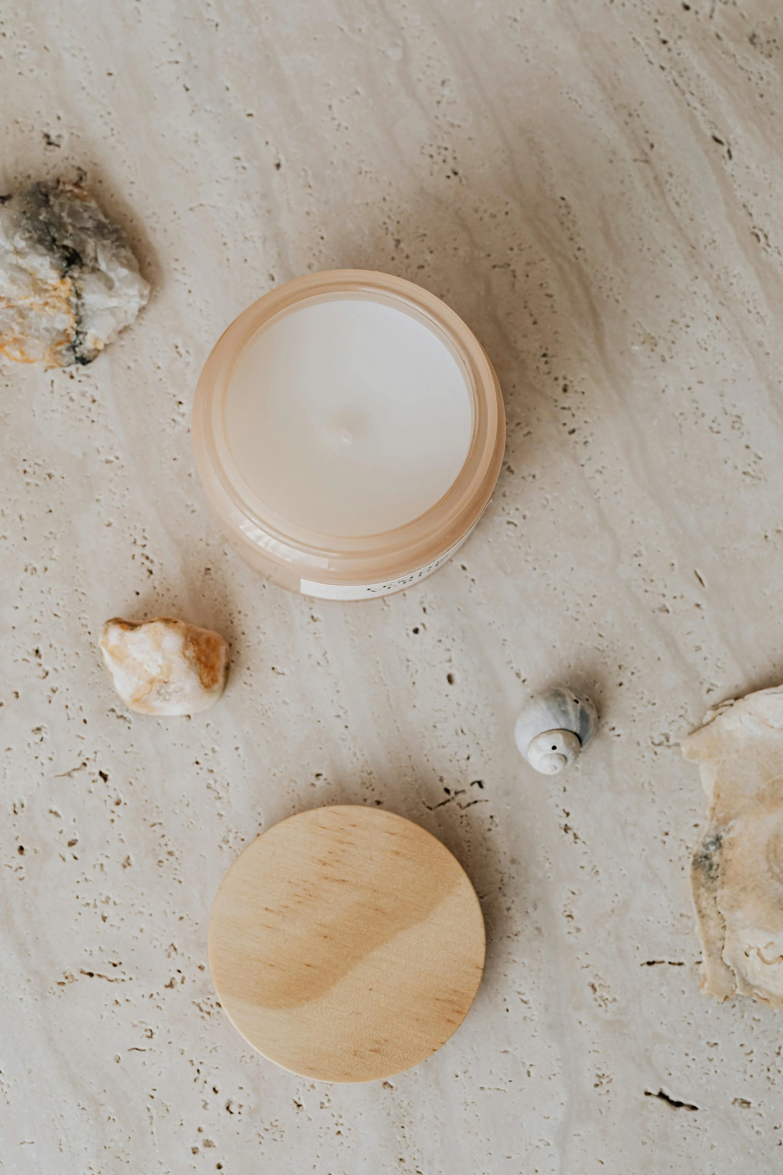 A jar of white cream or lotion with a beige lid, placed on a beige marble surface along with seashells and rocks.