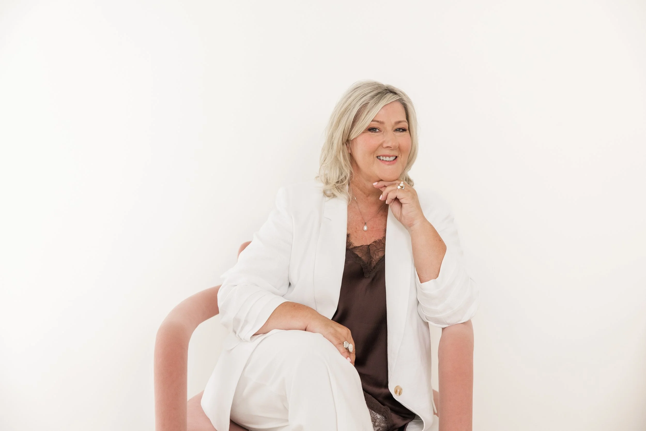 A smiling woman with blonde hair, wearing a white blazer and dark blouse, sitting on a pink chair against a white background.