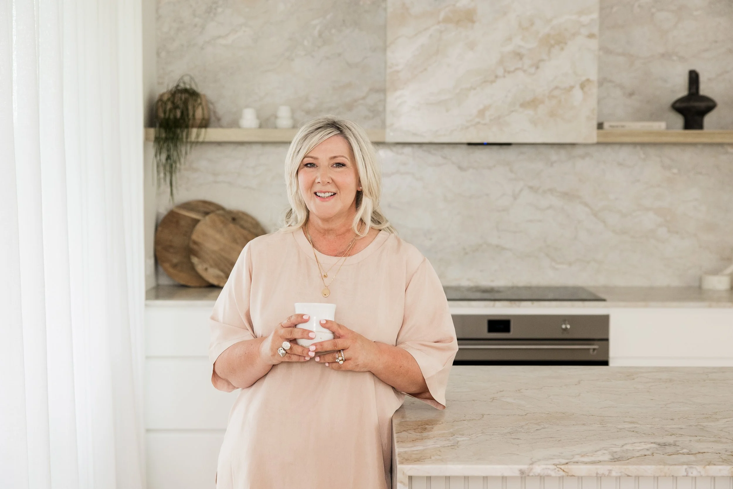 A woman with blonde hair smiling and holding a white mug in a modern kitchen with light-colored countertops and minimal decor.