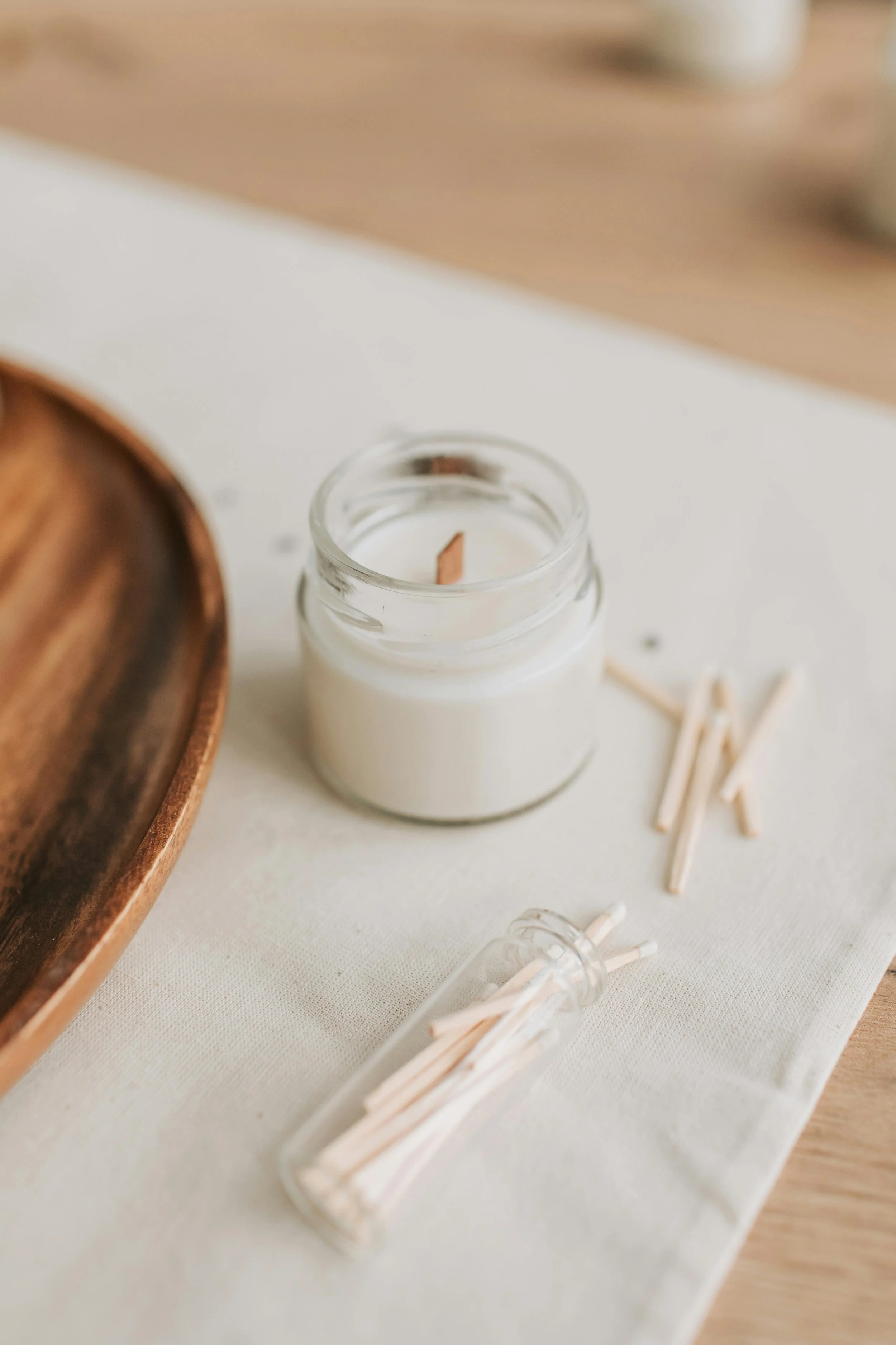 Close-up of a white candle in a glass jar with a wooden wick, placed on a white cloth, with some wooden stir sticks and a glass tube holding more stir sticks nearby, on a wooden surface.