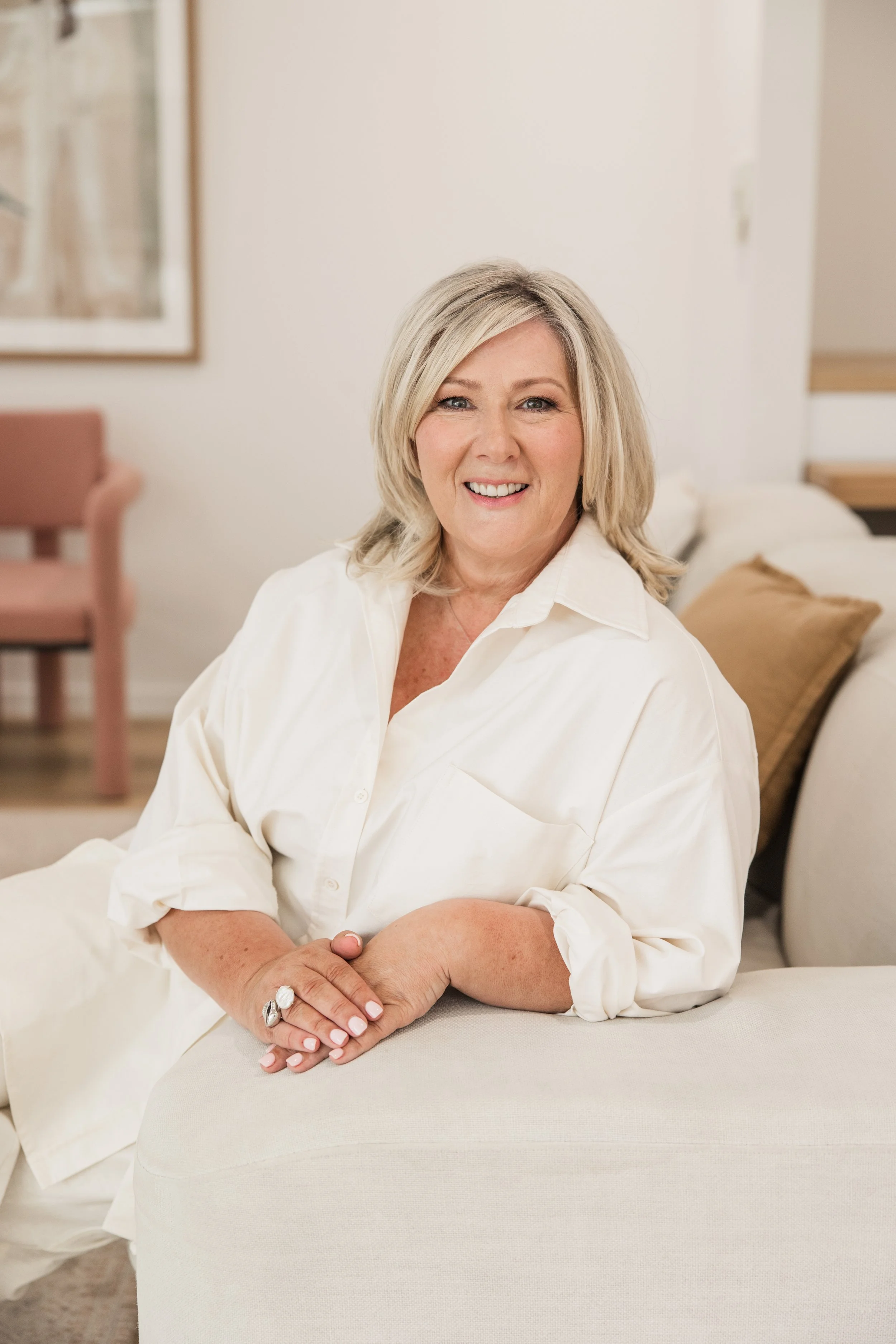 A smiling middle-aged woman with blonde hair sitting on a cream-colored sofa in a cozy, well-decorated living room