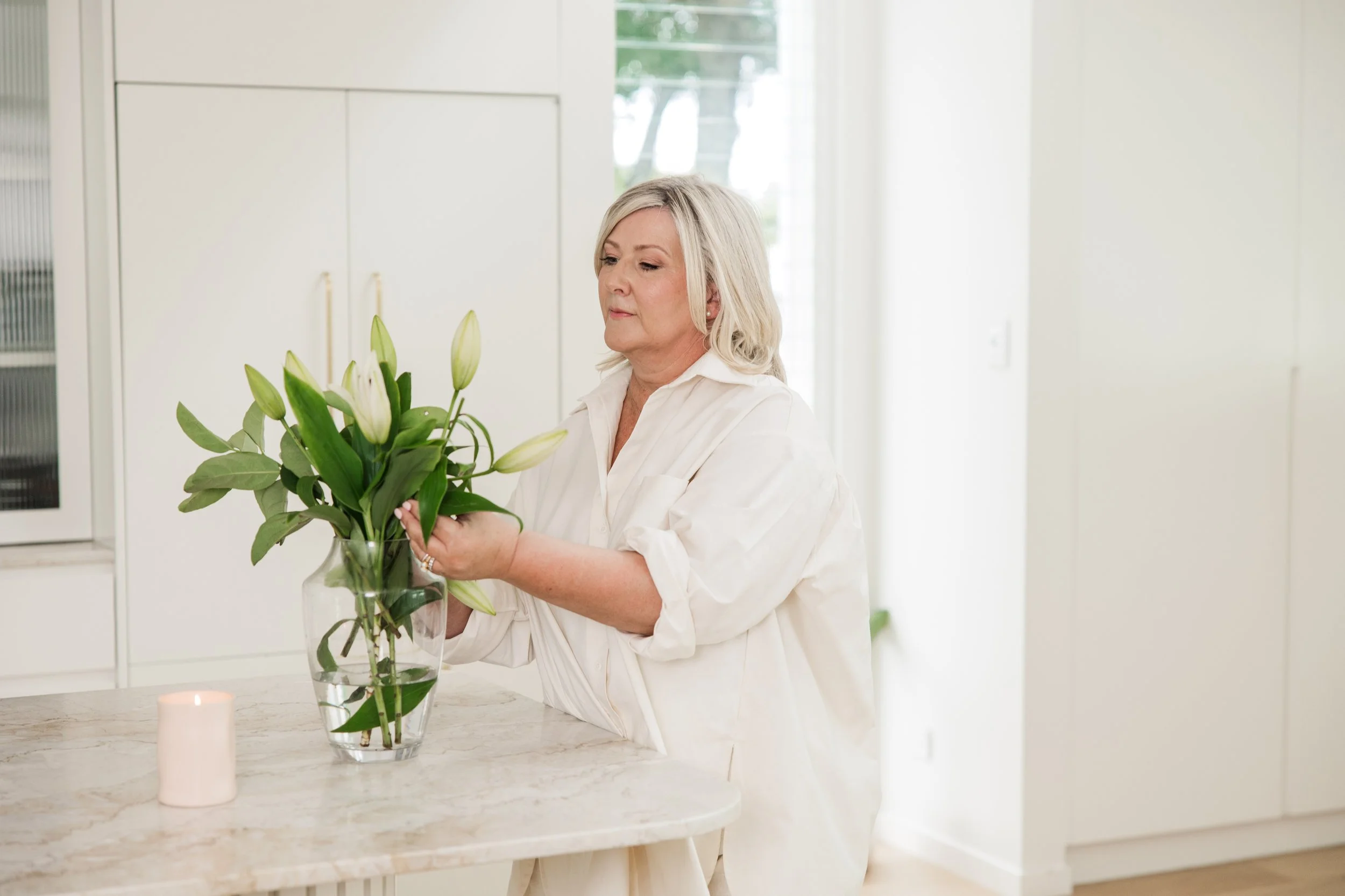 A woman arranging white lilies and green foliage in a glass vase on a kitchen counter, with a lit candle nearby.