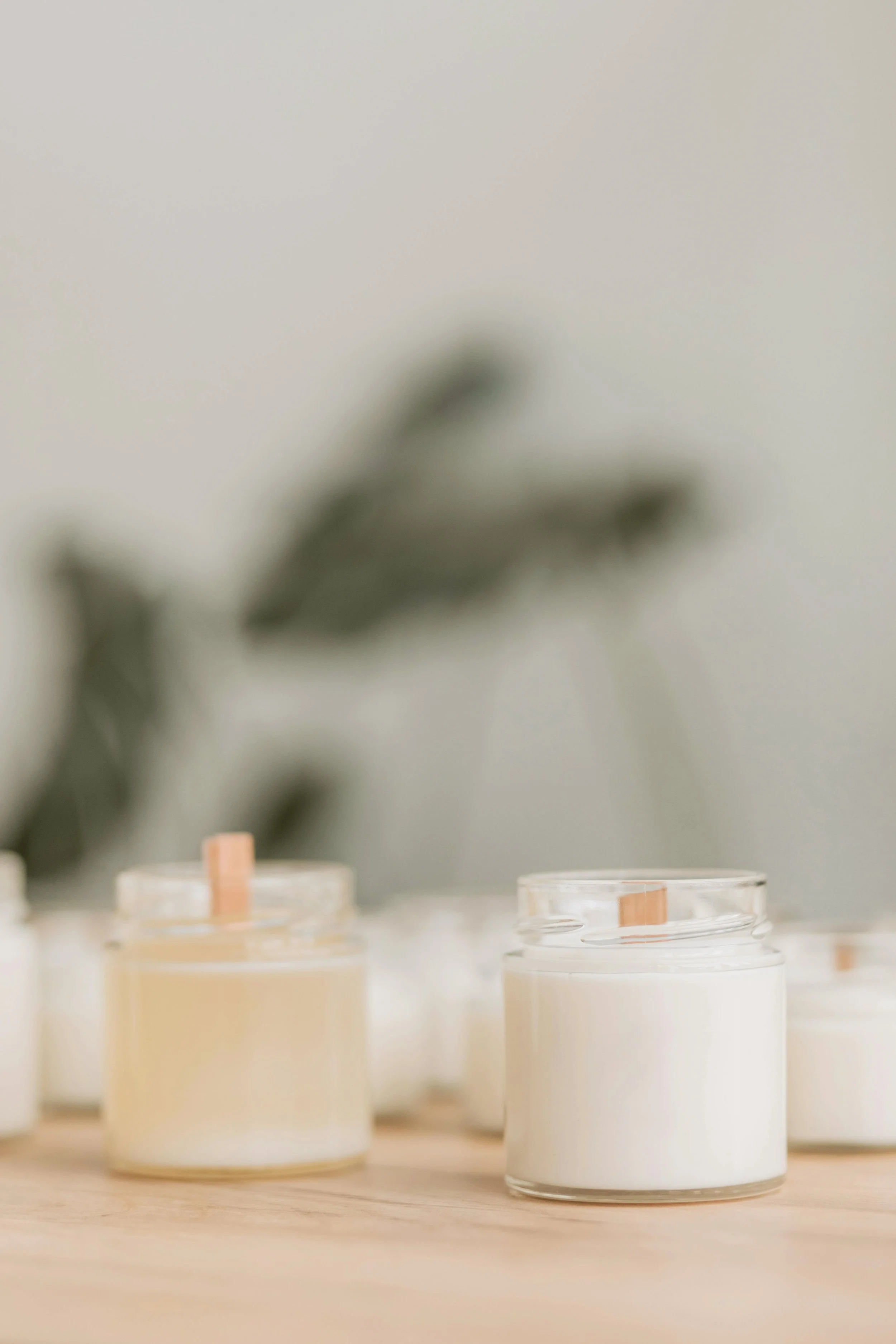 Several small candles in glass containers placed on a wooden surface, with blurred leaves or plants in the background.