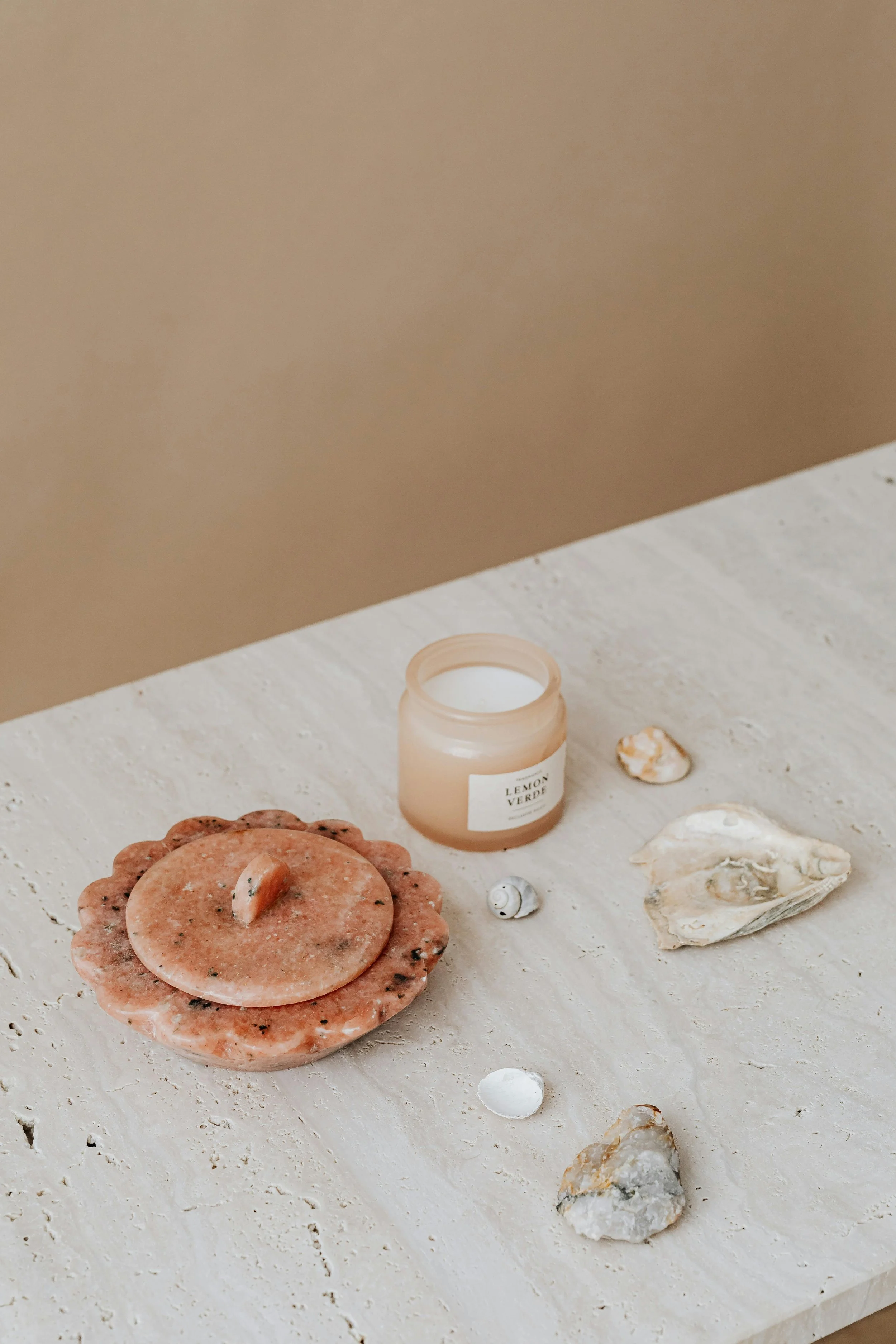 A collection of decorative items on a light-colored textured surface, including a small jar of candle labeled 'Lemon Verde,' seashells, and two pieces of salt rock.