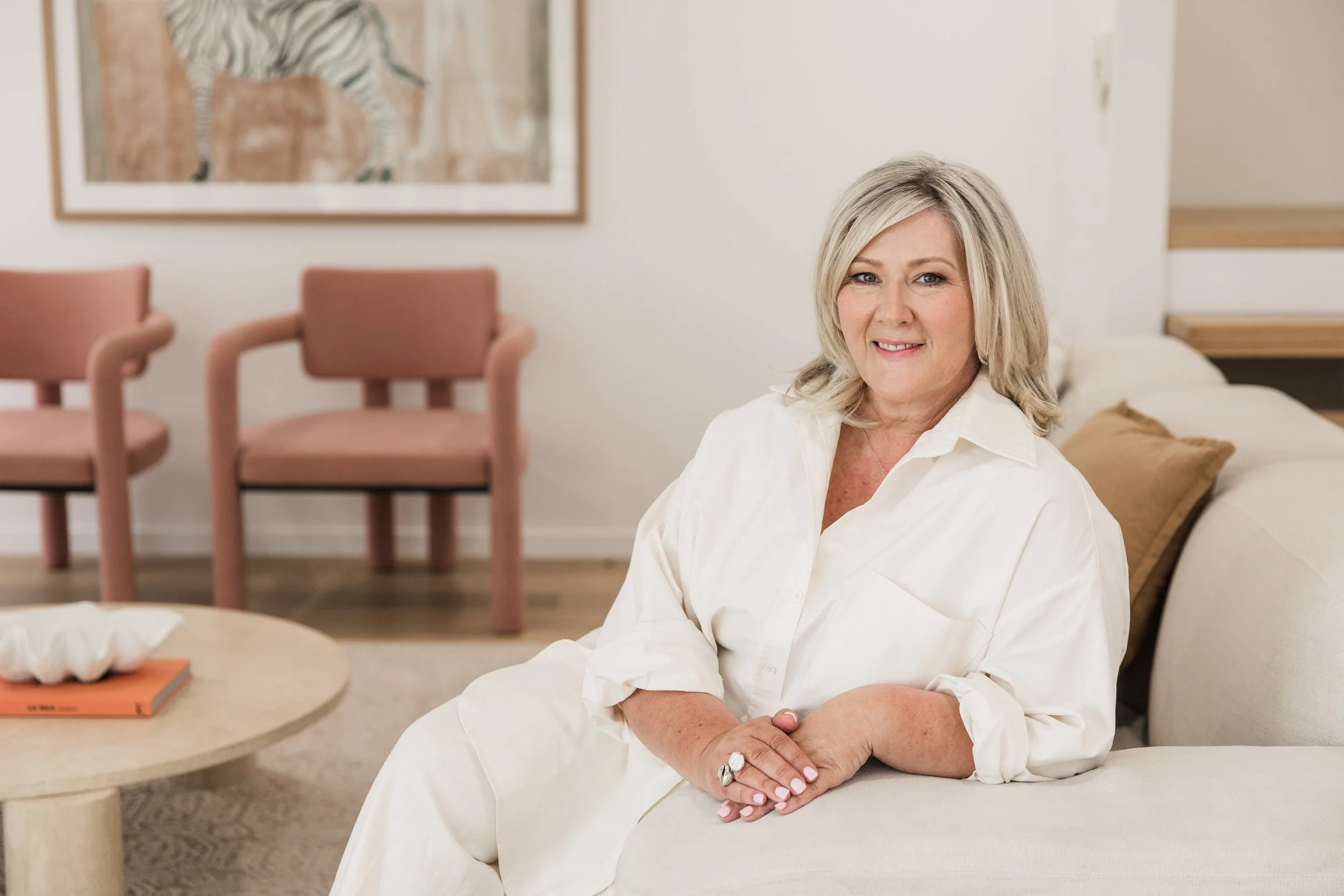A woman with shoulder-length blonde hair, wearing a white button-up shirt, sitting on a light-colored sofa.