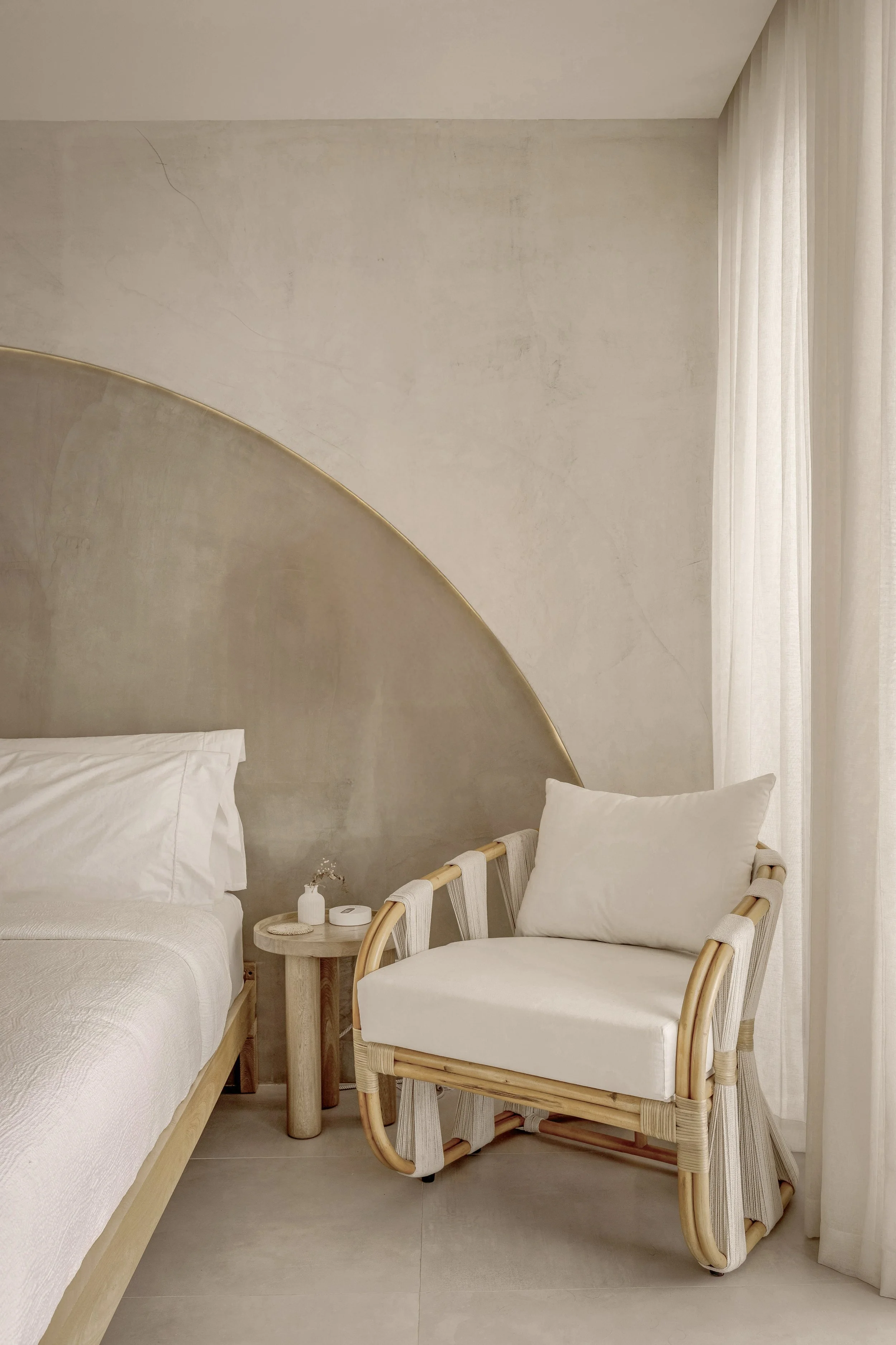 A minimalist bedroom corner with a bed, a small round side table, an armchair with a cushion, and cream curtains, all in neutral tones.