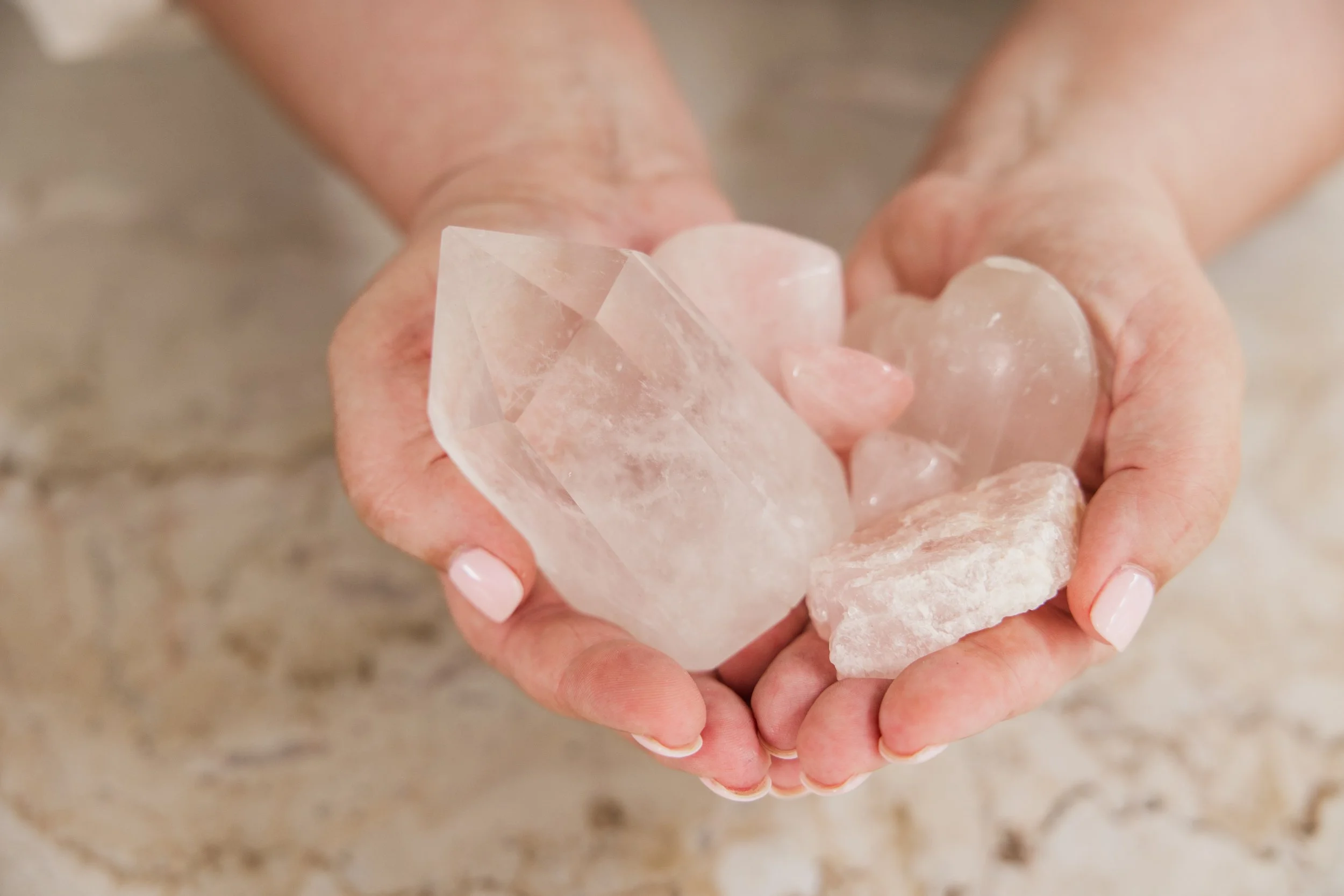 Hands holding various pink and white crystals, including heart-shaped and large rough pieces.