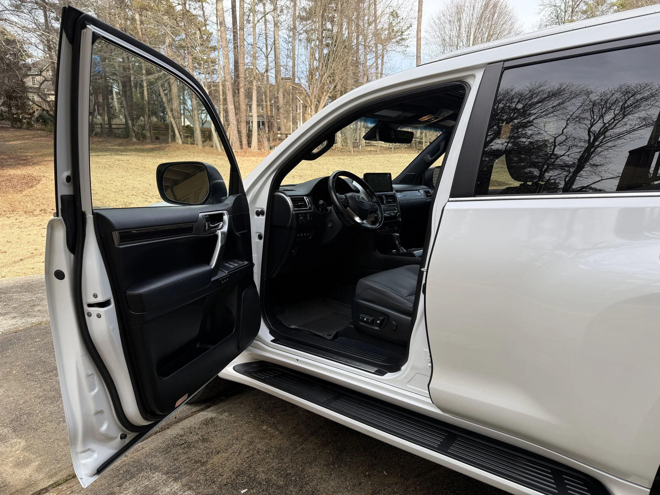 Open driver's side door of a white SUV, showing black interior with dashboard, steering wheel, and front seat, outdoors on a paved surface with trees and grass in the background.