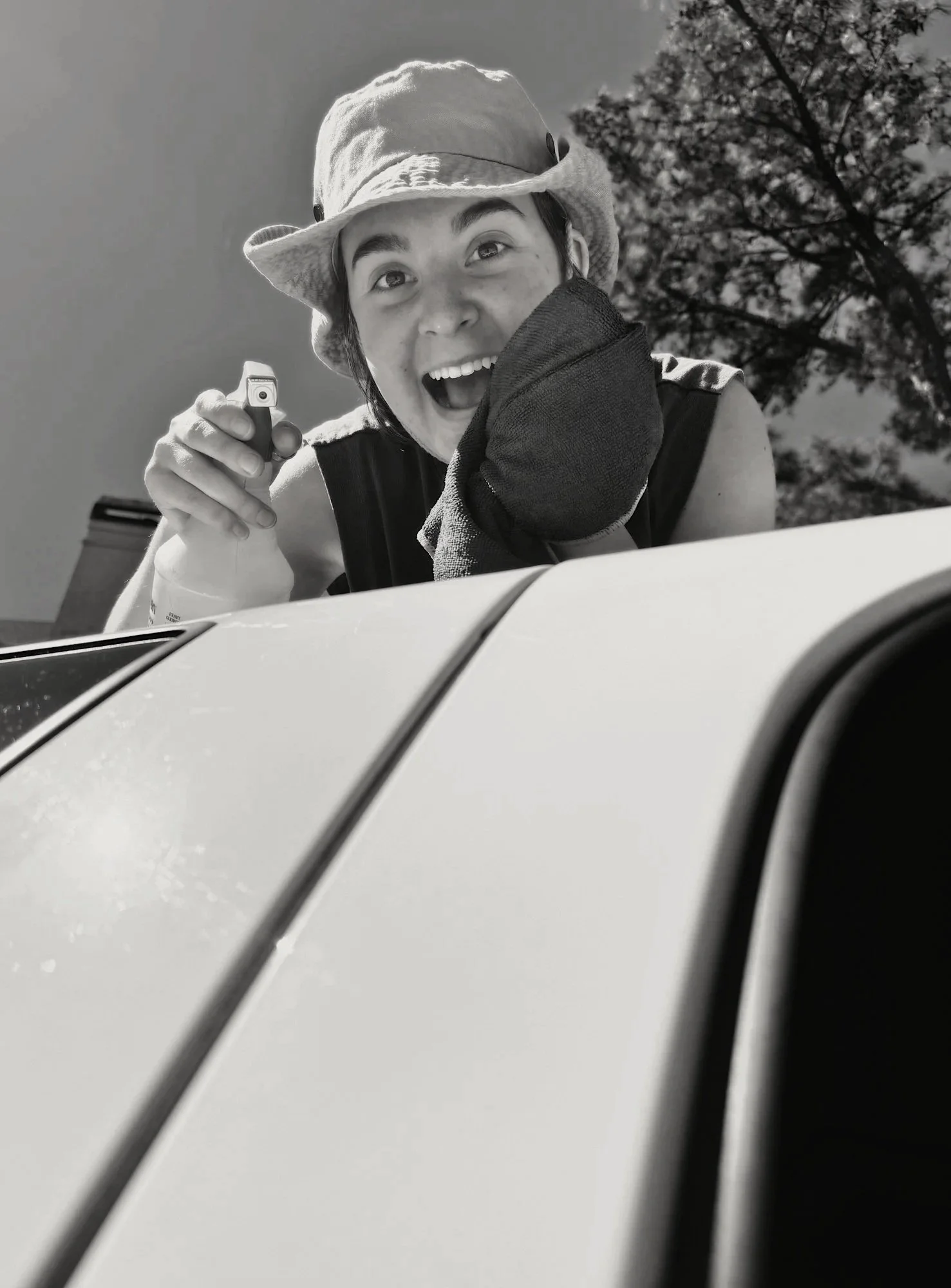 A person with a joyful expression is leaning over the hood of a car, holding a small object, possibly a camera or remote, and wearing a hat and a dark sleeveless top. The background shows trees and the sky.