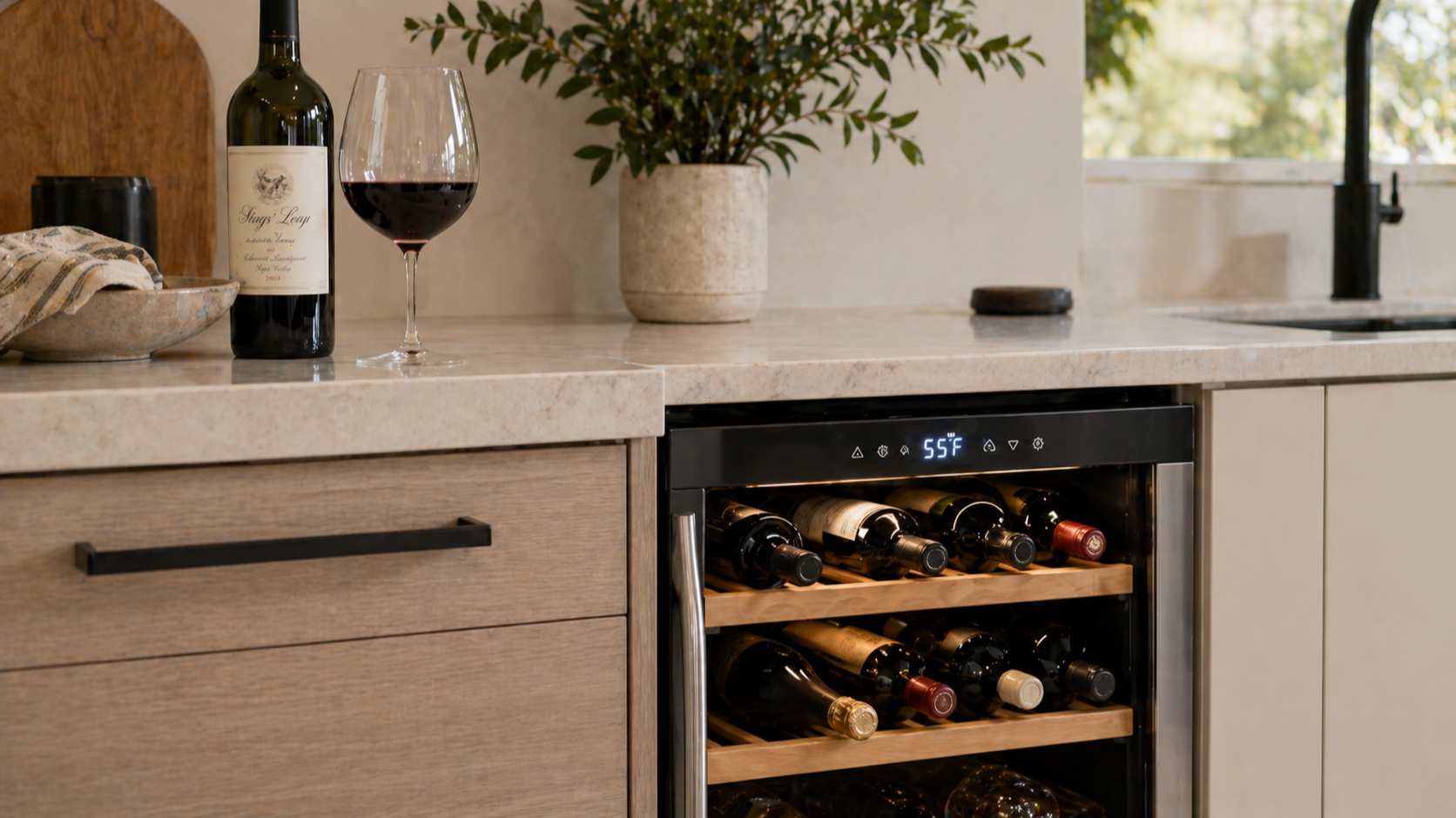 Modern kitchen with a built-in under-counter wine fridge beneath a light stone countertop, holding a mix of wine bottle shapes with slightly varied spacing; a single bottle and glass of red wine sit on the counter above in soft natural daylight.