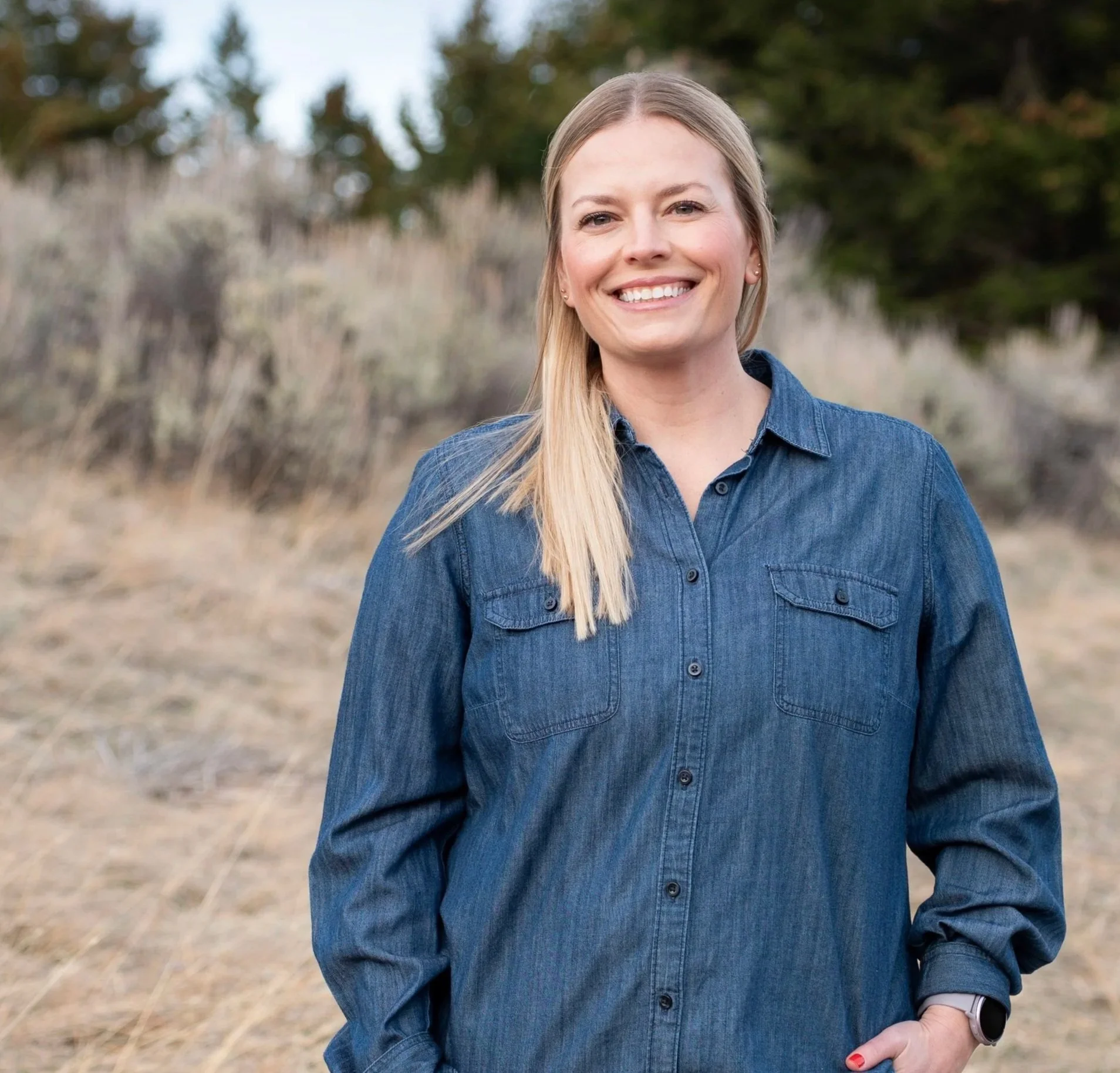 Smile of a young woman with long blonde hair, wearing a denim shirt, outdoors with blurred trees and dry grass in background.