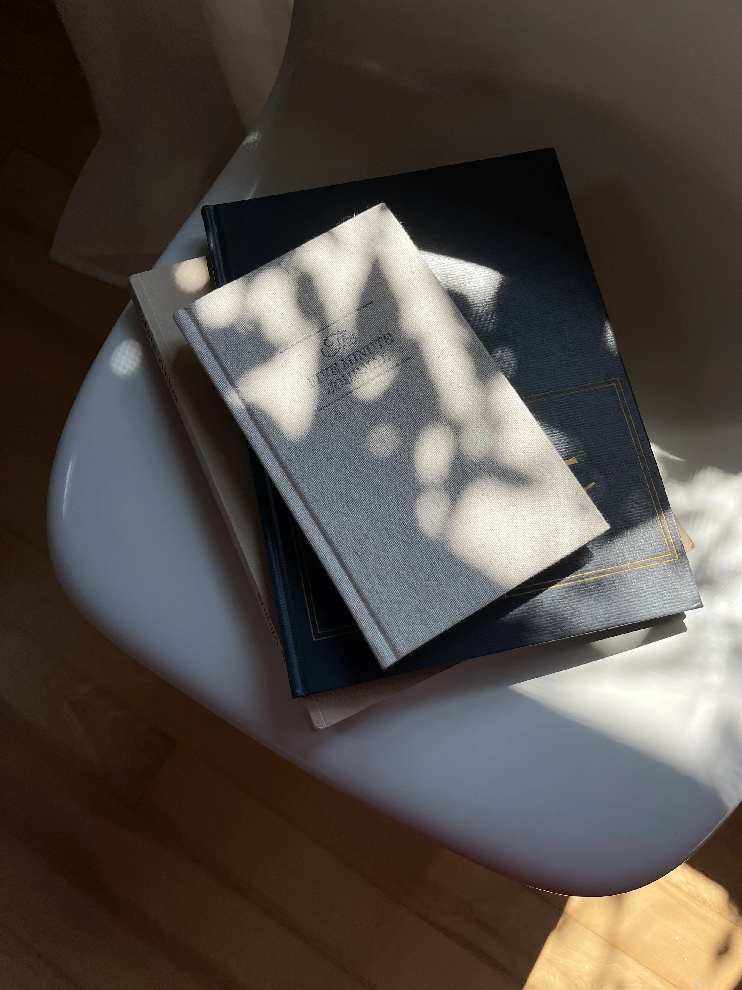 A stack of books on a white surface, with sunlight casting shadows of leaves on the top book.