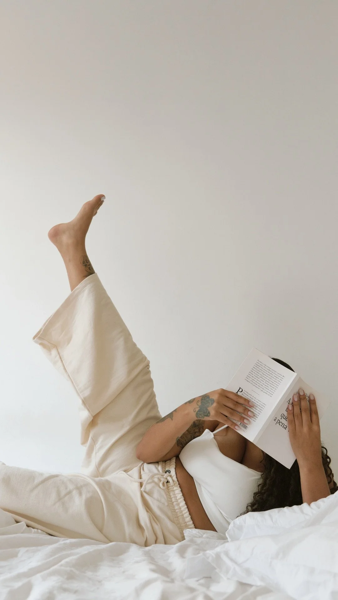 A woman lying on a bed, reading a book, with one leg raised in the air, wearing beige pajamas, with tattoos visible on her arm and thigh, in a minimalist room with white walls.