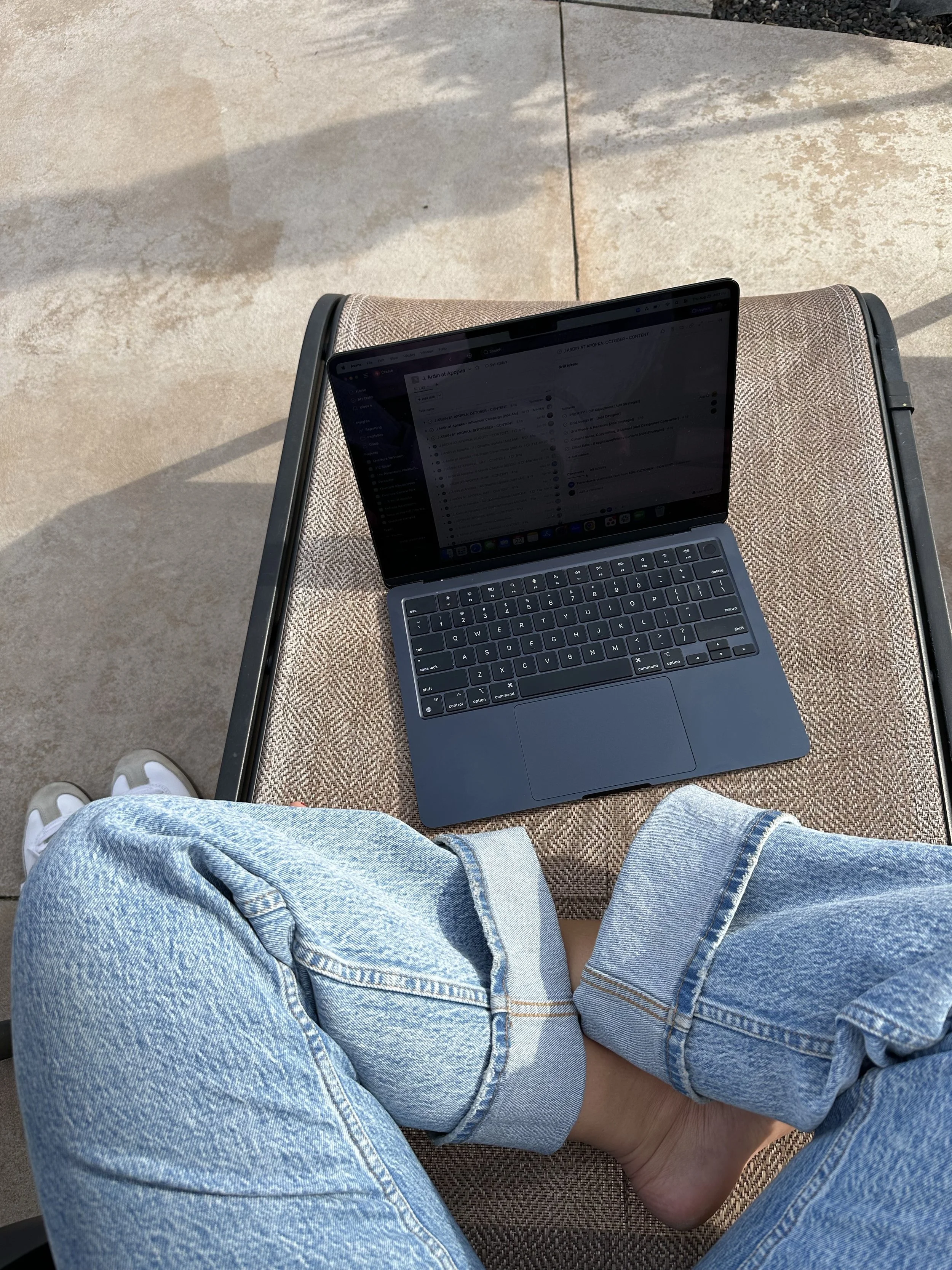A person sitting outdoors on a lounge chair with their legs crossed, wearing ripped blue jeans and white sneakers, with a laptop on their lap, on a concrete surface.