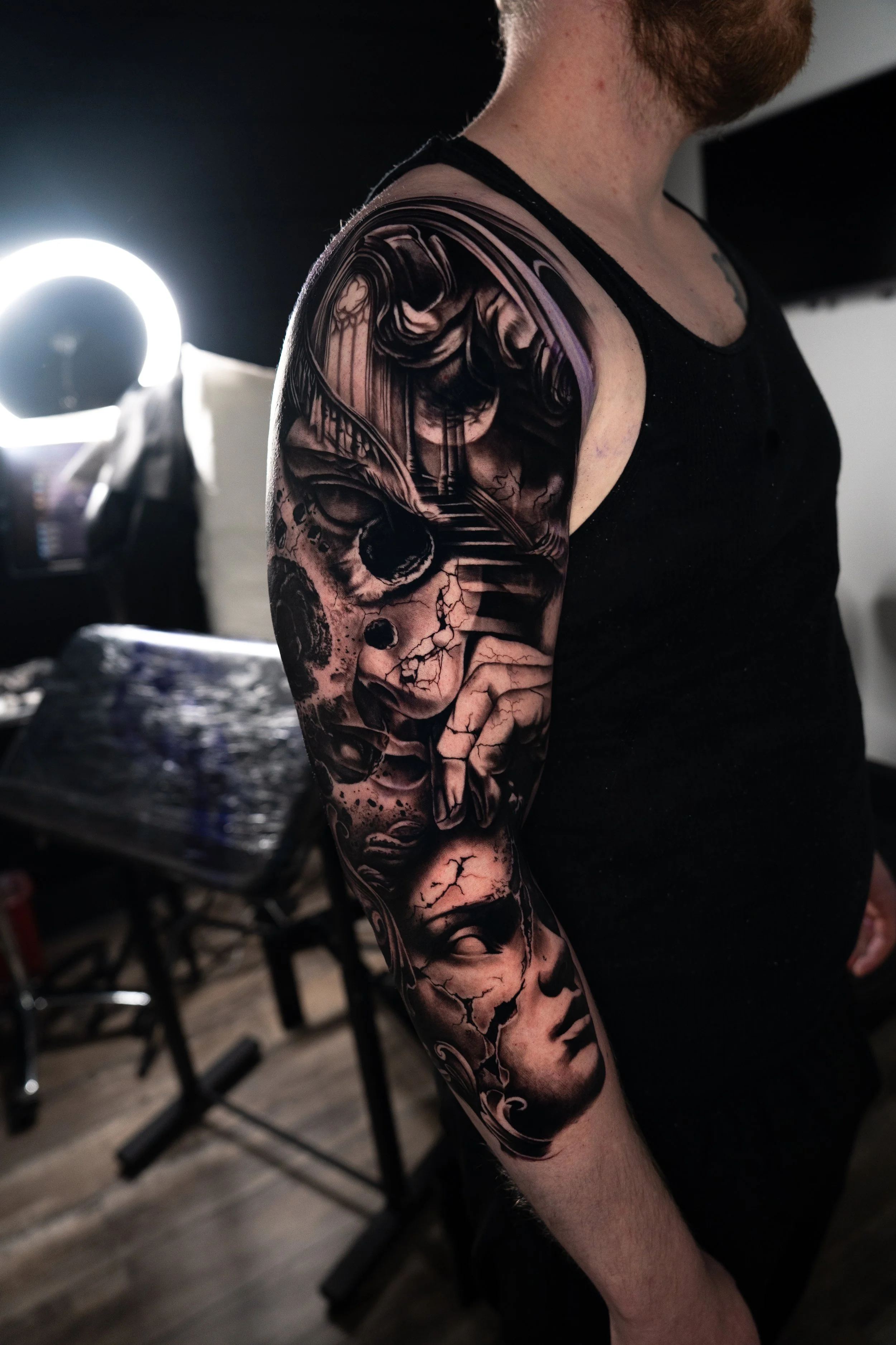 Close-up of a person's arm with a detailed black and gray tattoo featuring a skull, cracked faces, and a classical sculpture face.