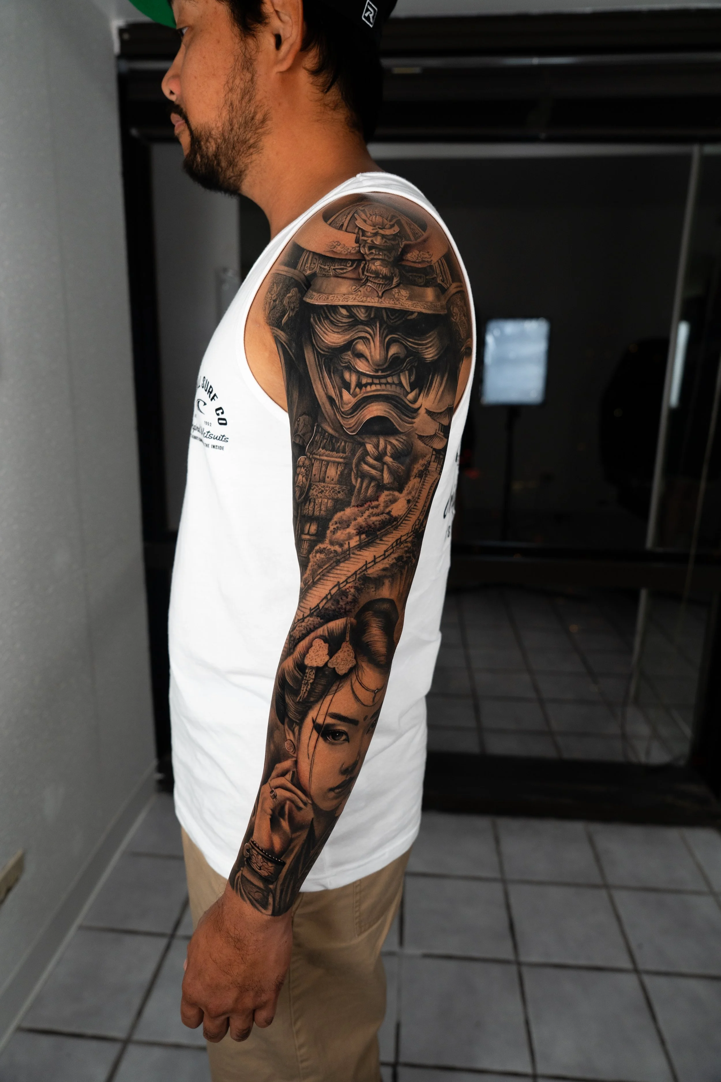 A man with a black beard and a tattoo sleeve on his right arm, wearing a white sleeveless shirt, standing indoors with dark background and tiled floor.