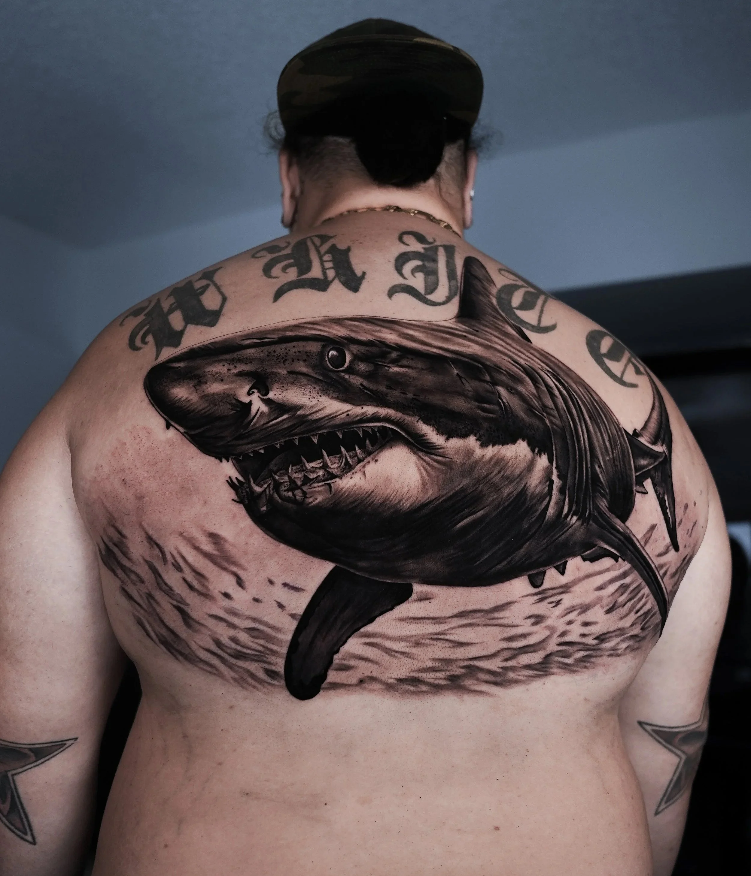 Tattoo of a great white shark on a man's back, with tattoos and a cap.