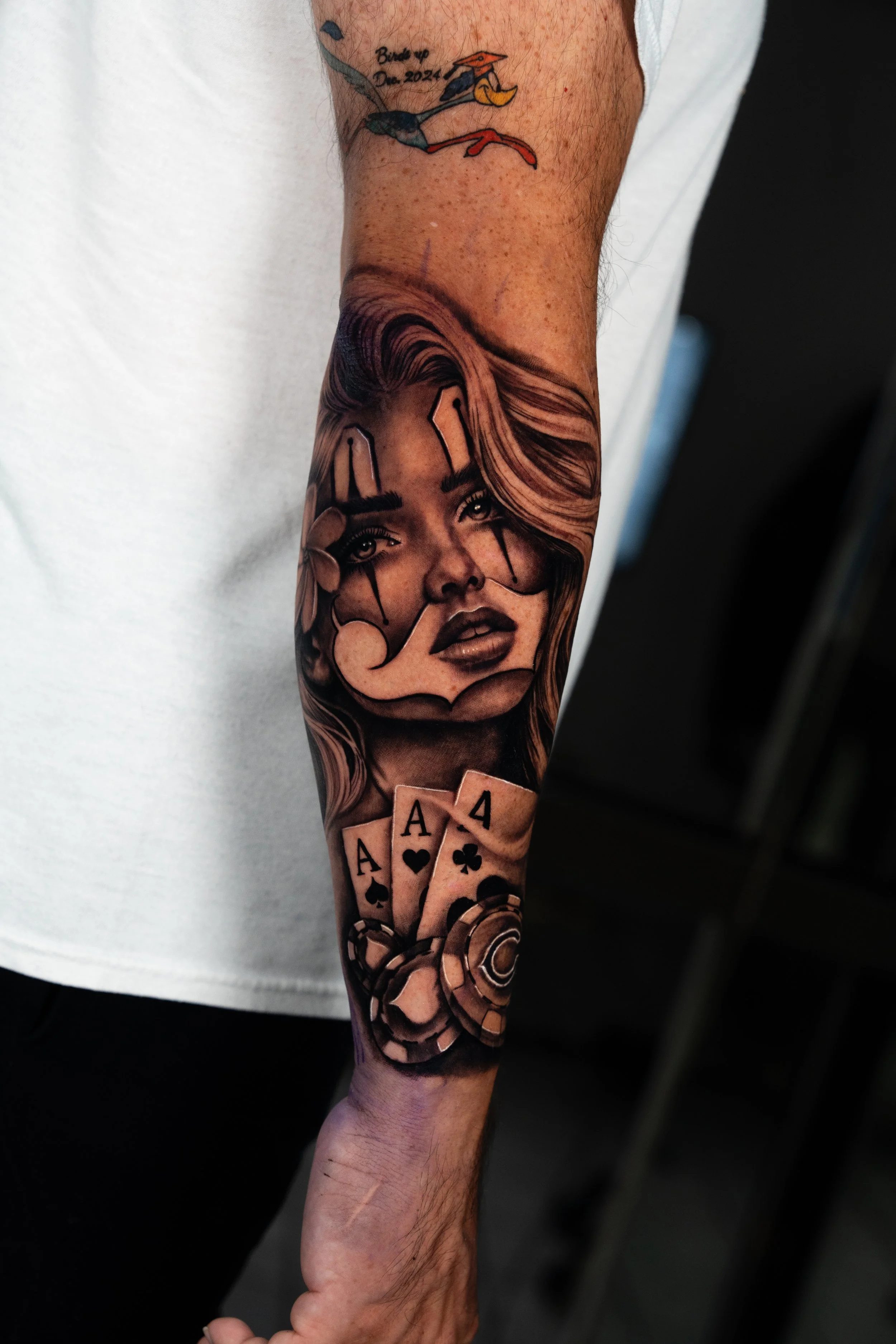 A black and gray tattoo of a woman's face with flowing hair, surrounded by poker chips and playing cards with four aces, on a person's forearm.