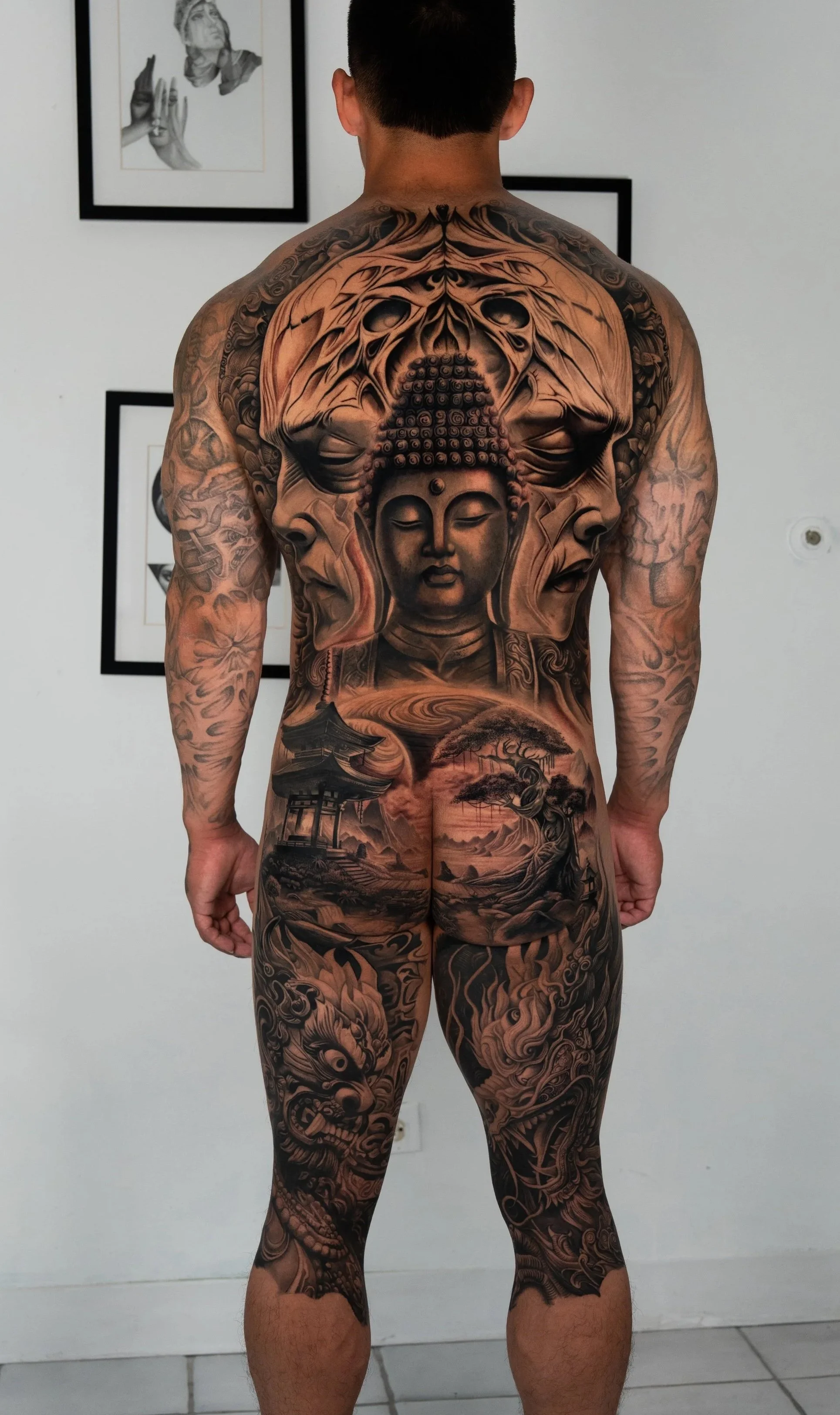 A shirtless man with extensive black and gray tattoos covering his back, arms, and legs, featuring a large Buddha face on his back, a pagoda, trees, and a dragon.
