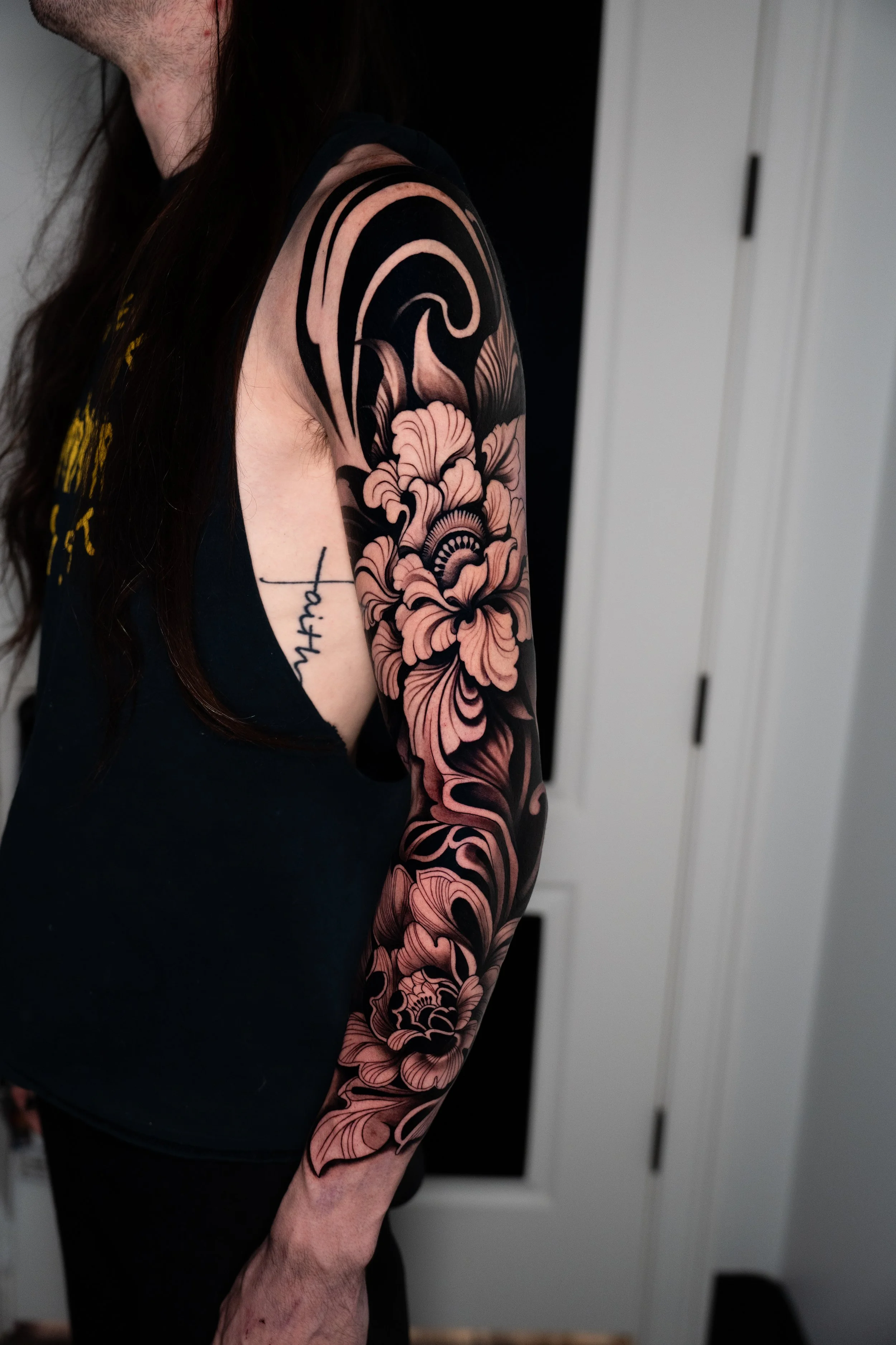 Close-up of a person's arm with a large black and pink floral tattoo extending from the shoulder to the wrist, with a portion of a black sleeveless shirt and a tattooed word visible on the side.