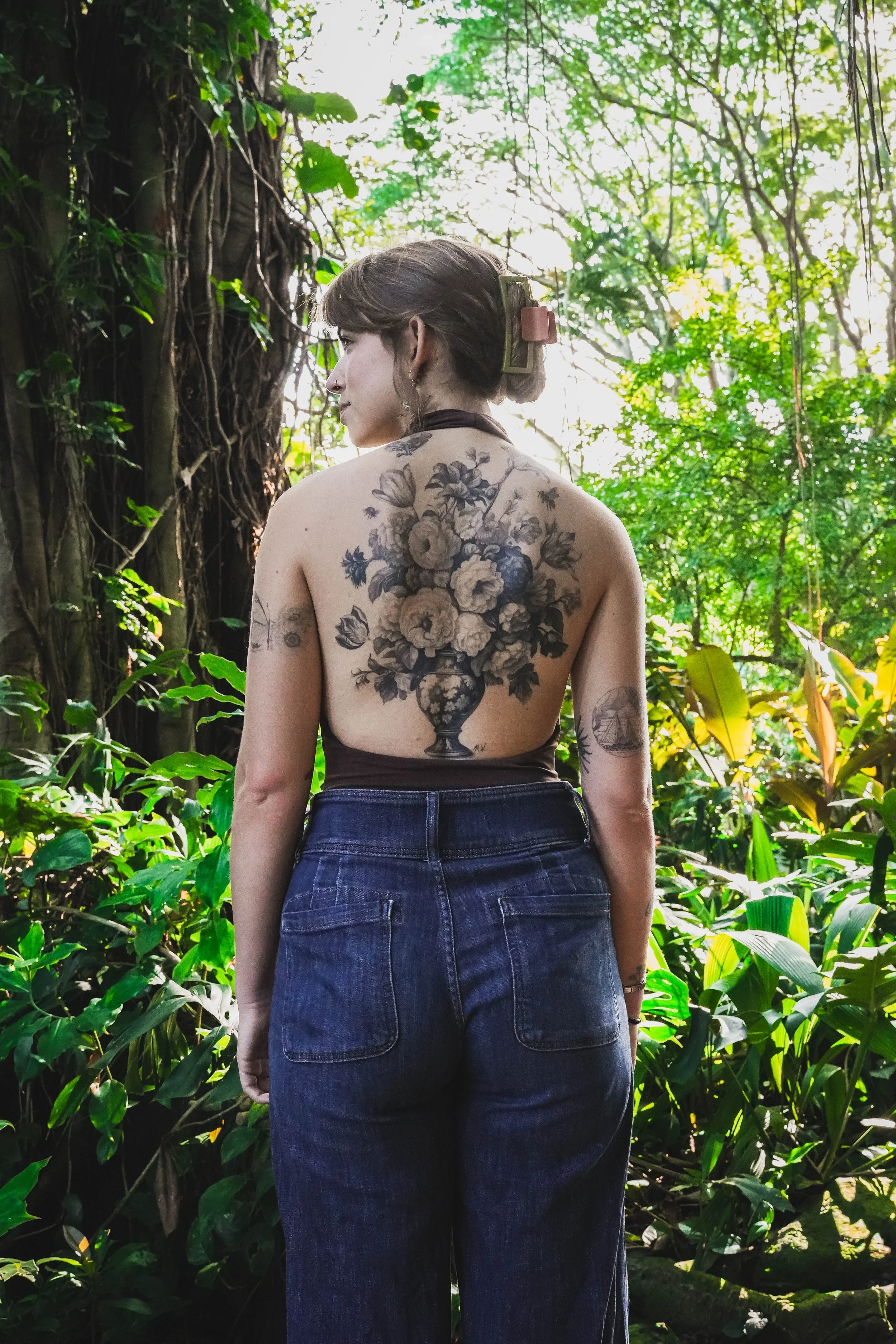 A woman standing in a lush, green jungle, seen from behind, with large floral tattoos on her back, wearing dark high-waisted jeans and a top with her hair held up.