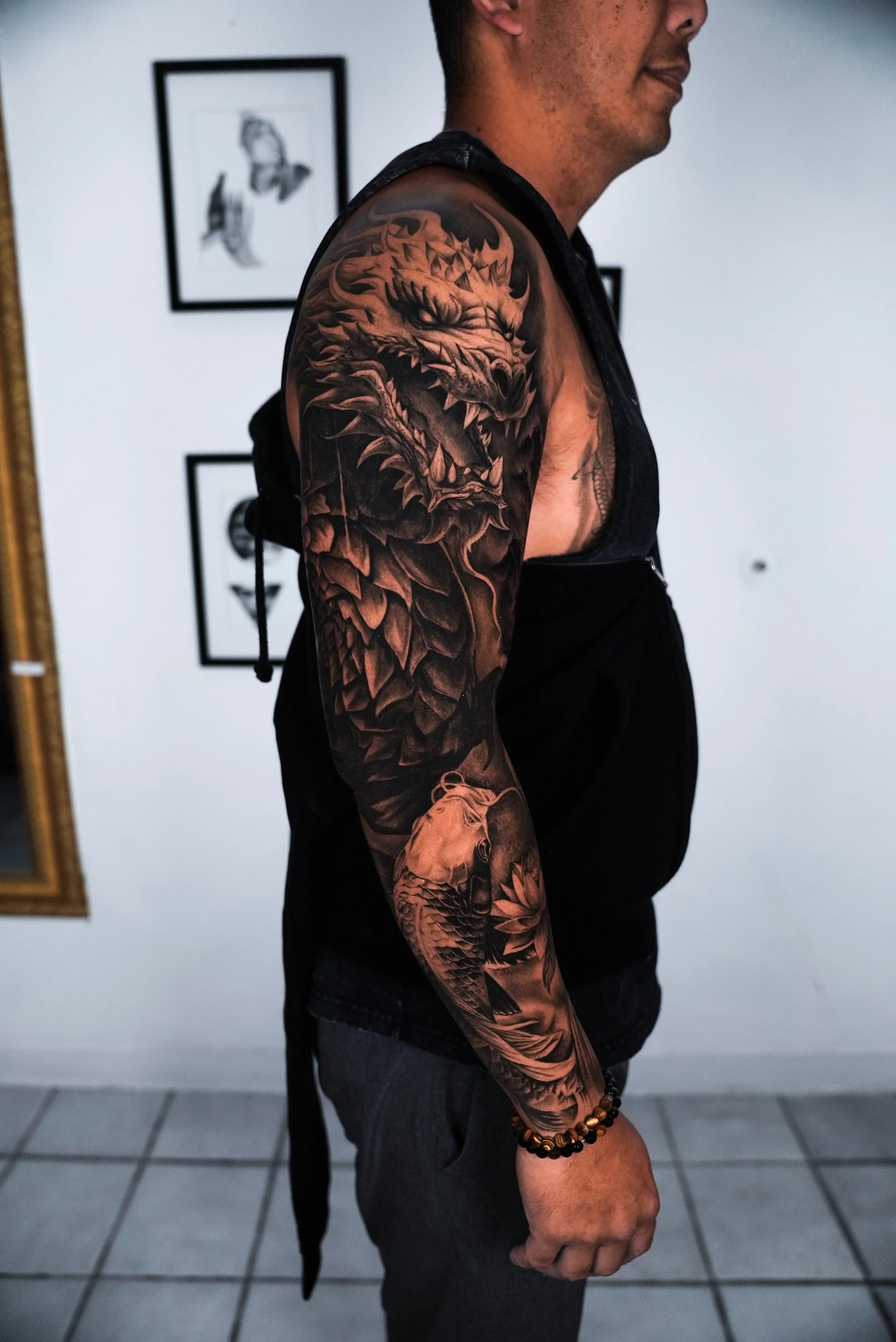 A man with a large, detailed black-and-gray dragon tattoo on his upper arm and a koi fish tattoo on his forearm, standing indoors against a white wall with framed artwork.