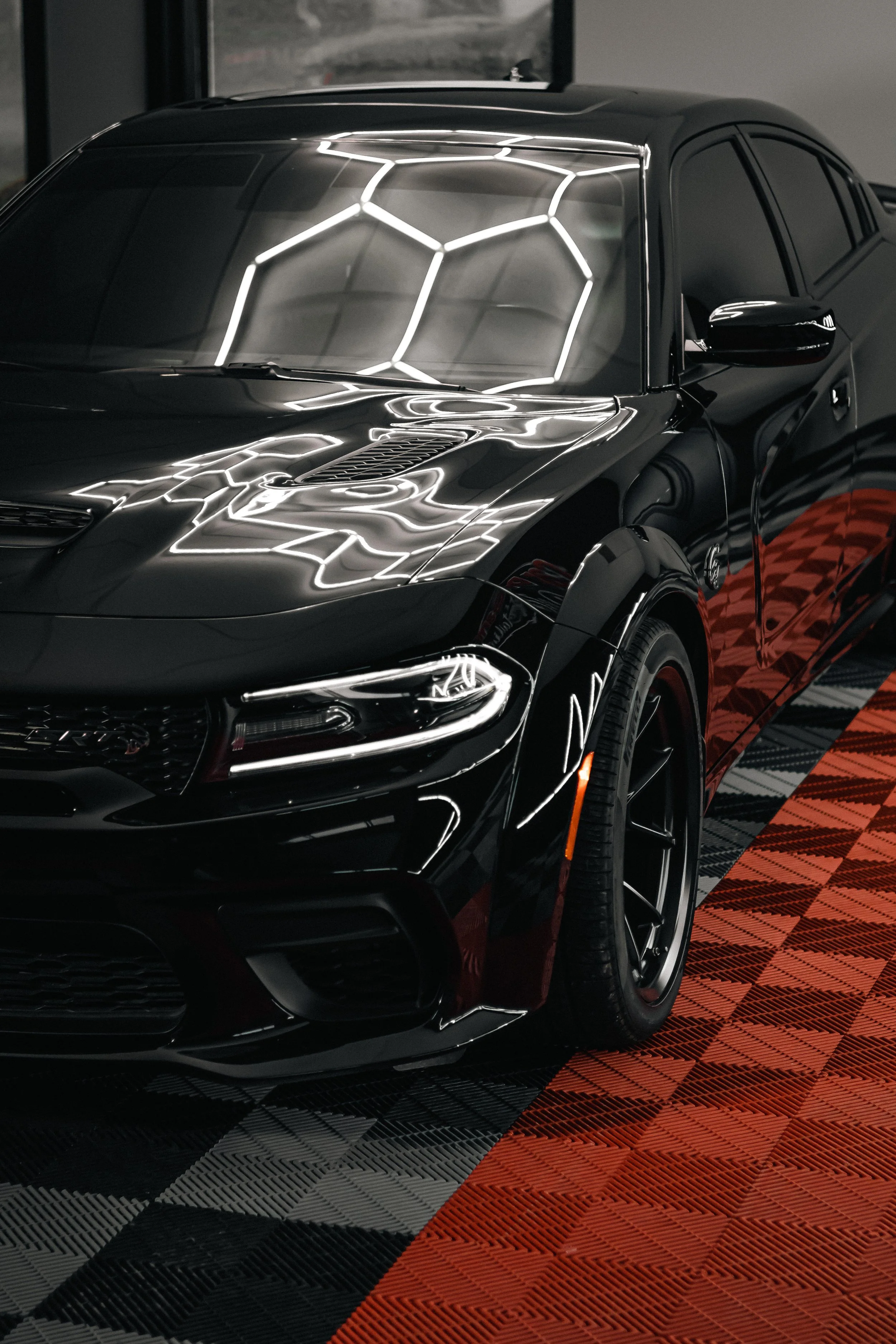 Black luxury sedan parked indoors, glossy surface reflecting bright white geometric LED lights, red and black patterned flooring underneath.