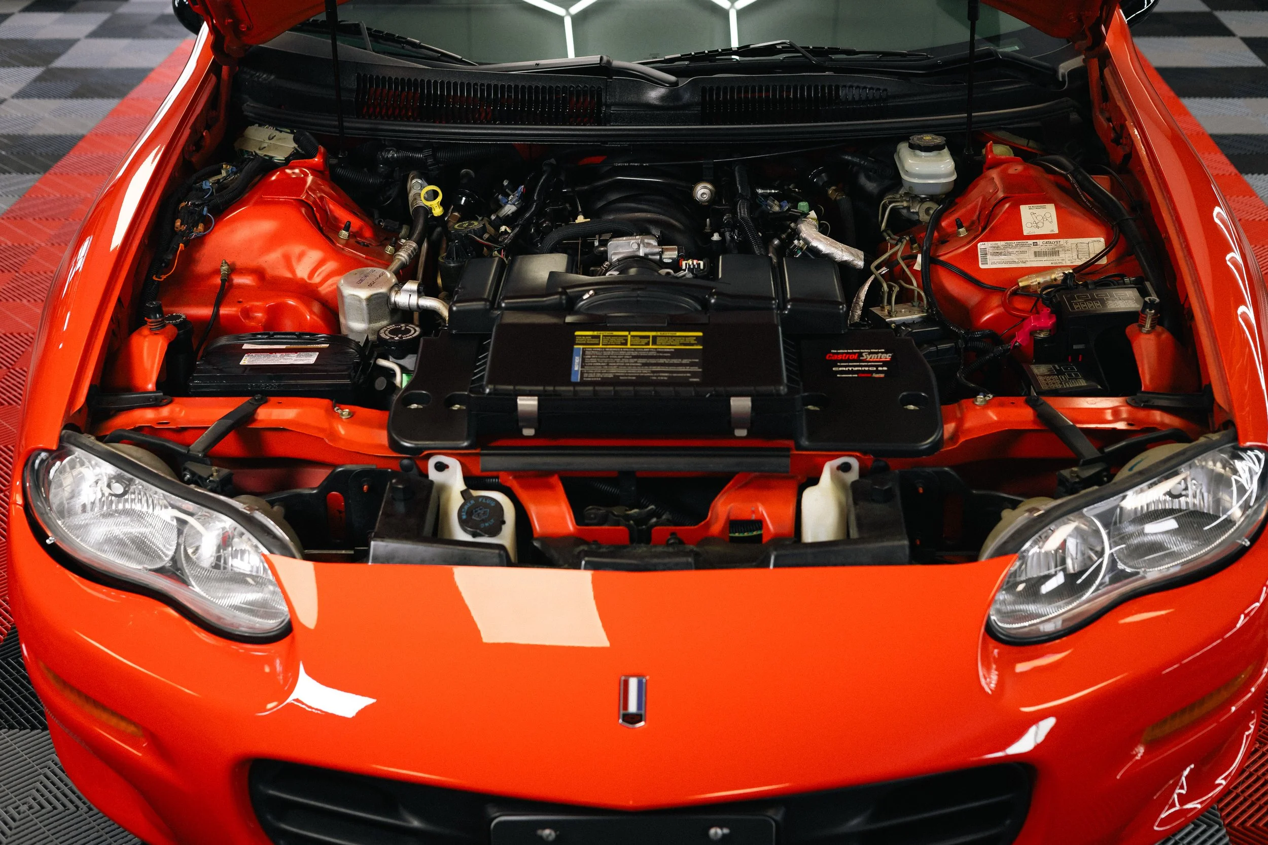 Front view of a red car with the hood open, revealing the engine bay. The car is parked indoors on a black and gray checkered floor.
