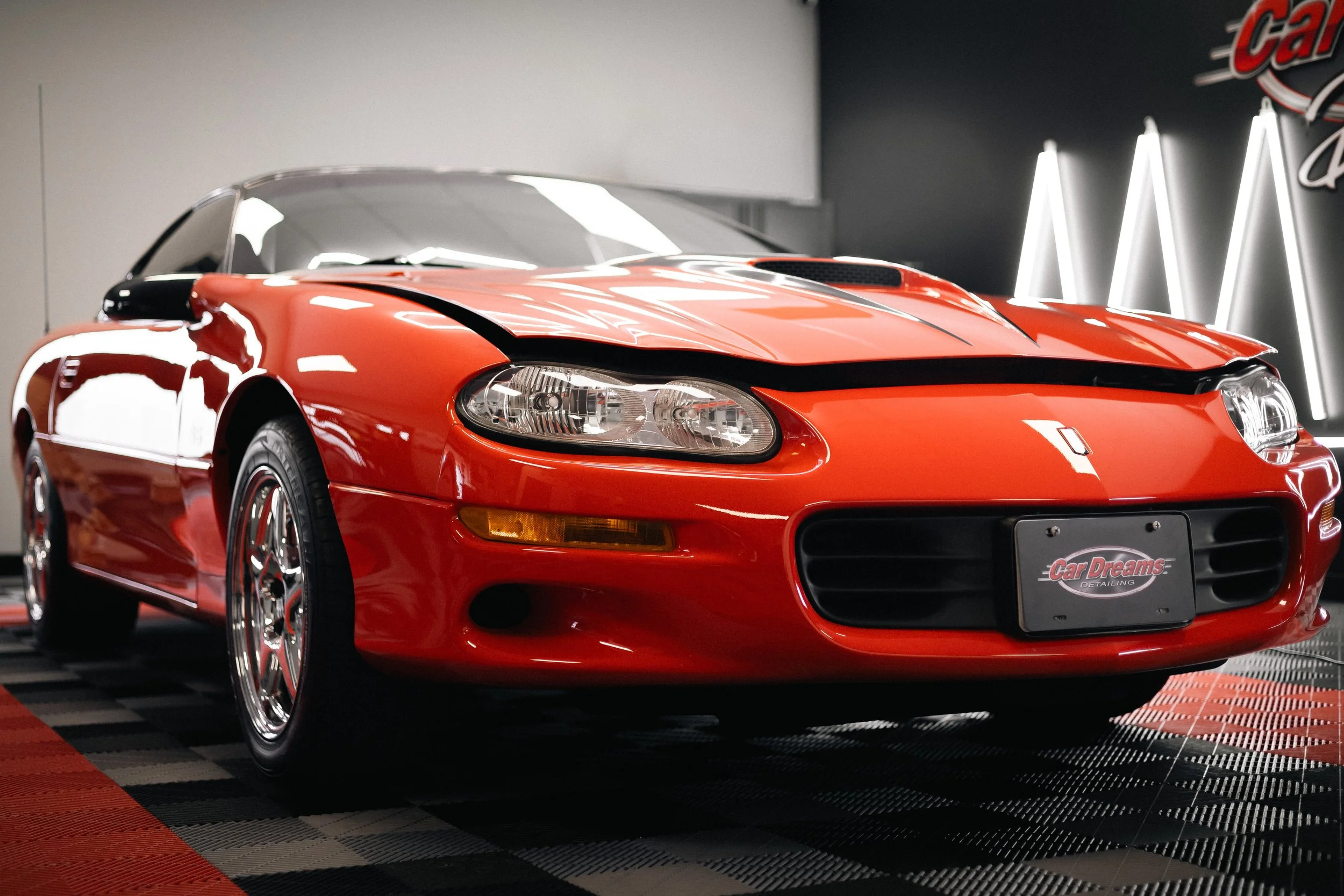 A red sports car with a sleek design, black detailing, and shiny chrome wheels, parked indoors against a dark wall with illuminated neon signs.