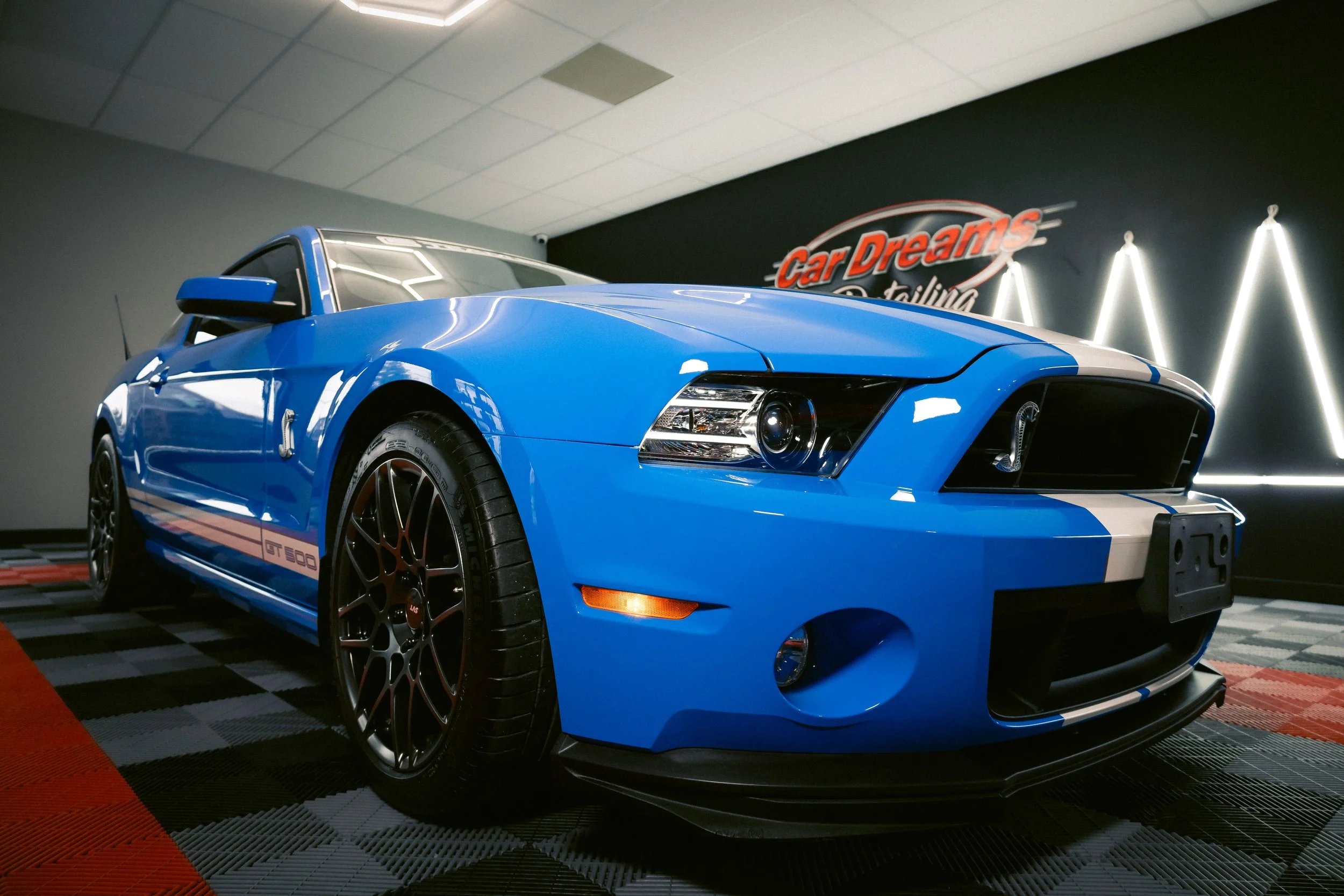 A blue Ford Mustang Shelby GT500 sports car inside a showroom with a black wall and neon sign reading 'Car Dreams detailing' in the background.