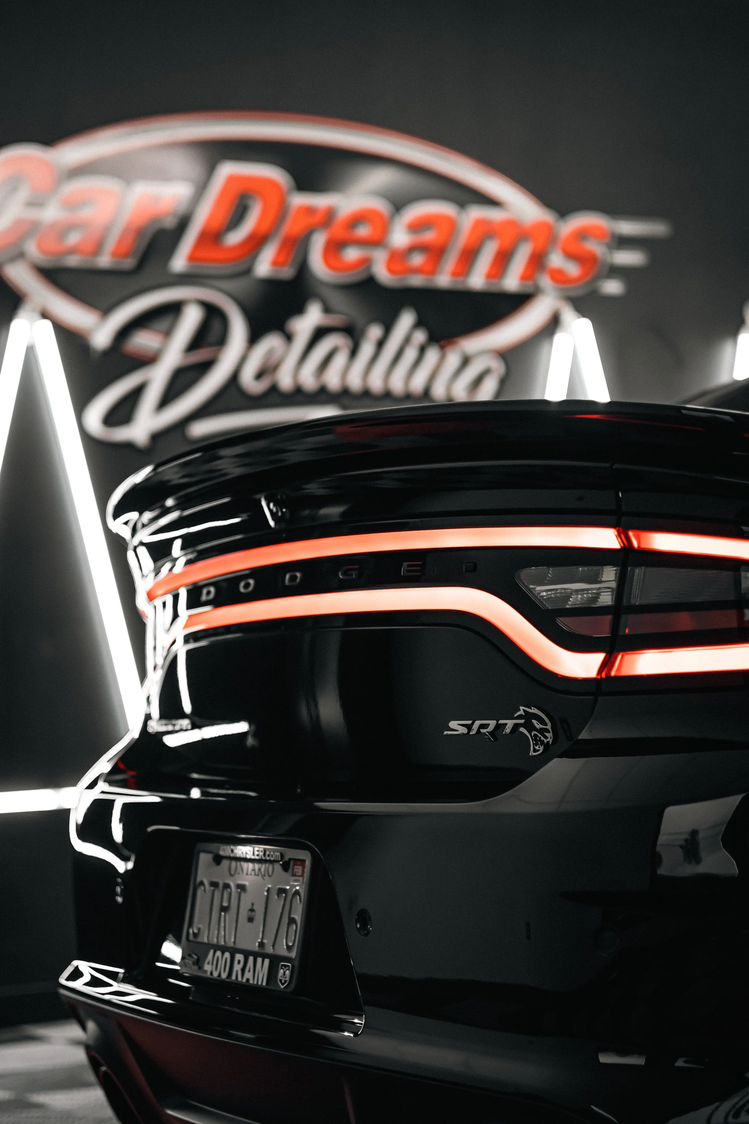 Rear view of a black Dodge Charger SRT Hellcat with illuminated taillights, showing part of a Nevada license plate, in a parking garage. In the background, a neon sign reads 'Car Dreams Detailing'.
