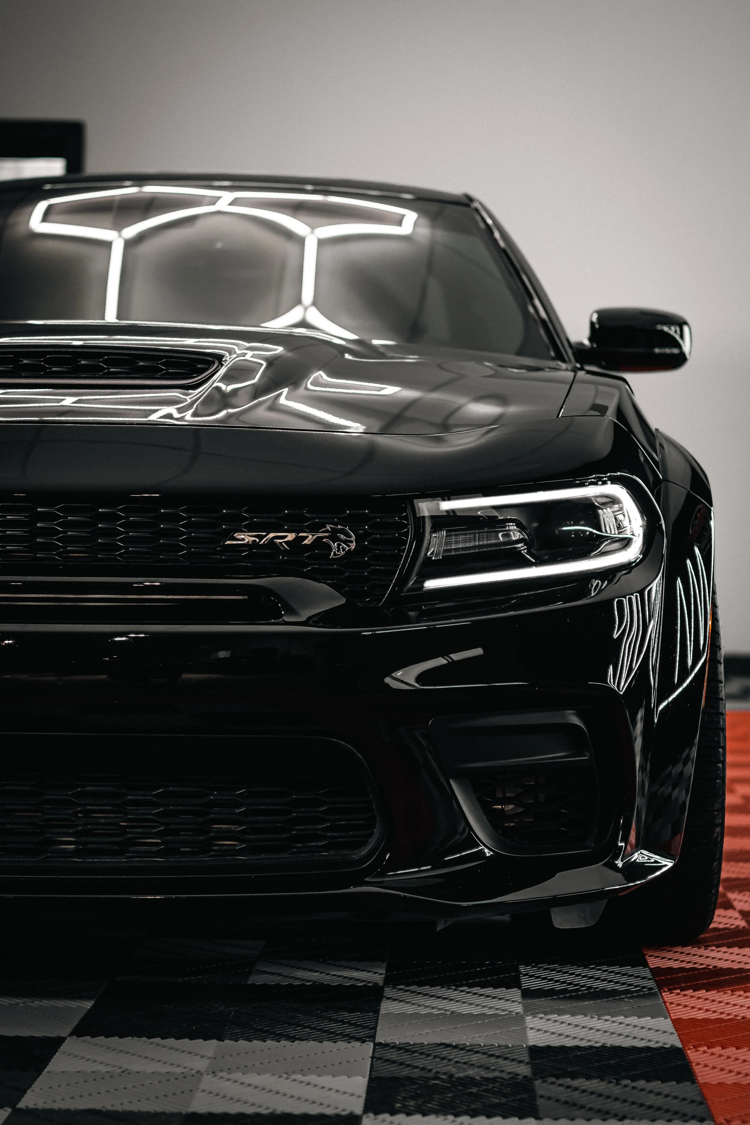 Close-up of a black sporty car with a sleek front grille and headlights, parked on a checkered floor with lighting reflections.