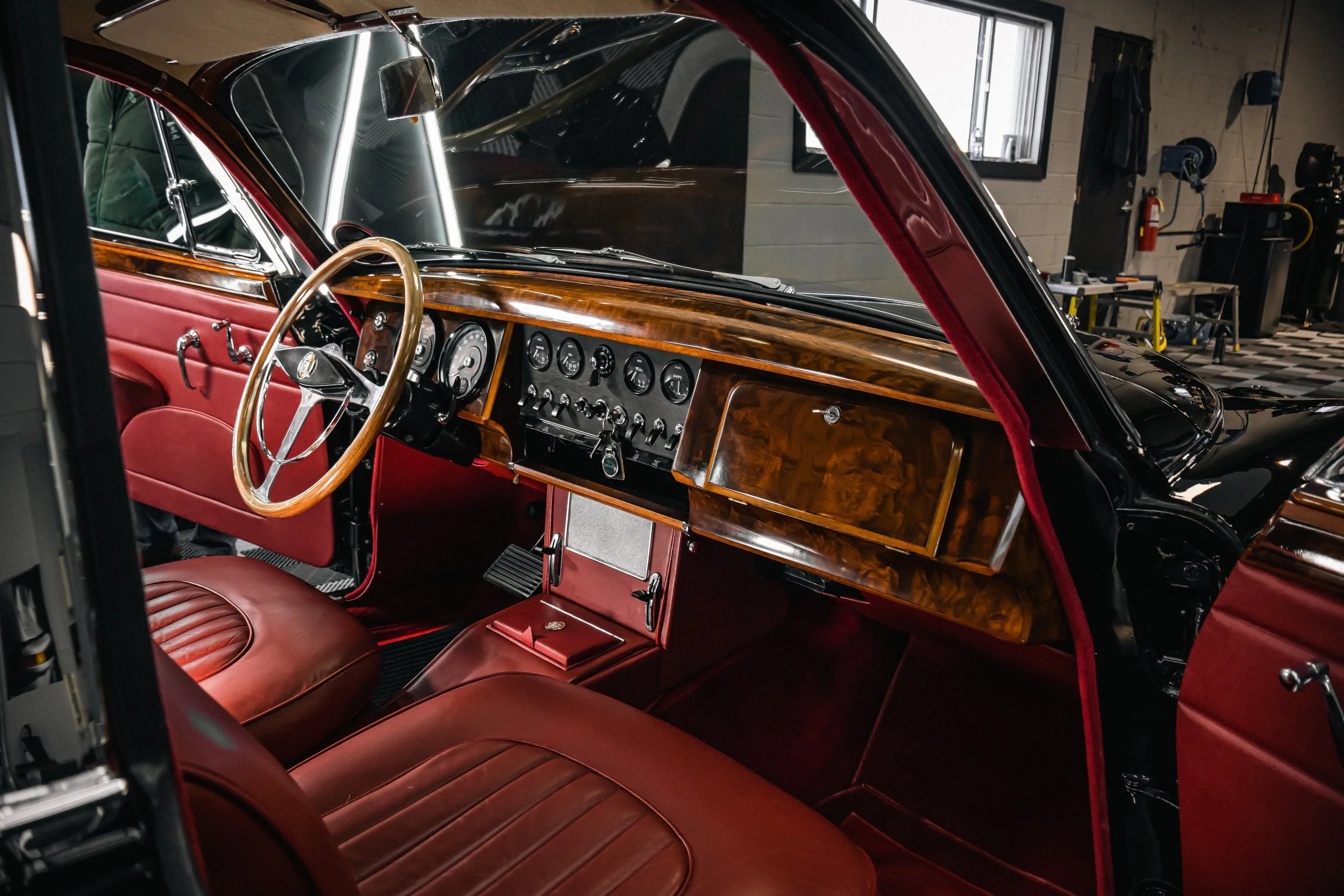 The interior of a vintage car featuring red leather seats, a woodgrain dashboard with multiple gauges, a wooden steering wheel, and a window view of a garage or workshop.