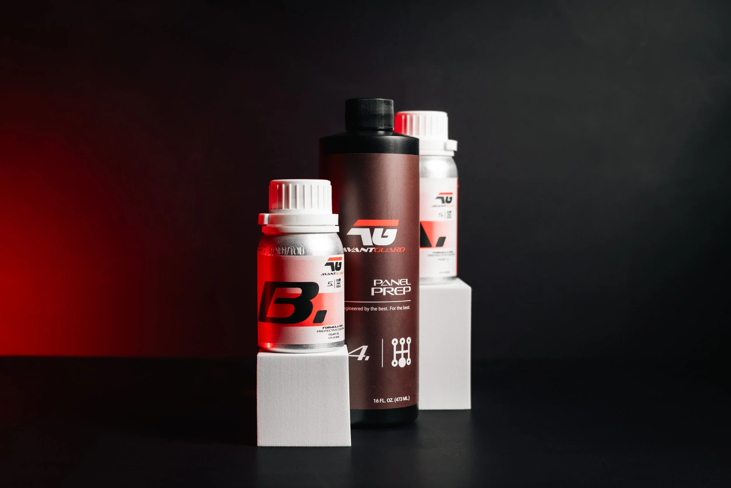 Three bottles with black, white, and red caps, and a black and red label, placed on white boxes against a dark background.