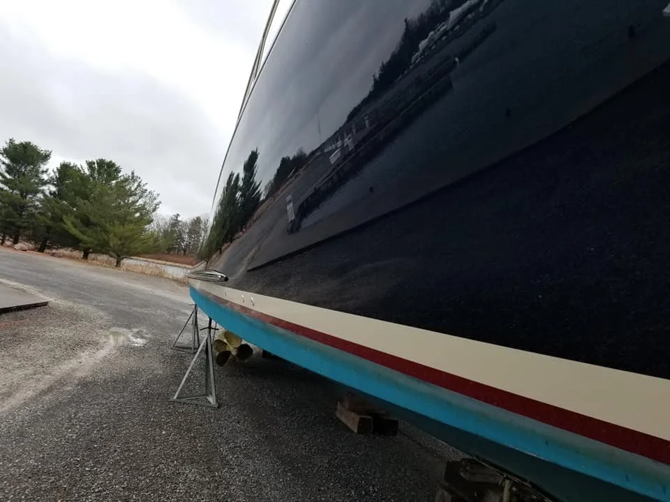 Photo of the side of a black boat on a trailer, parked on a gravel area. Some trees and a cloudy sky are visible in the background.