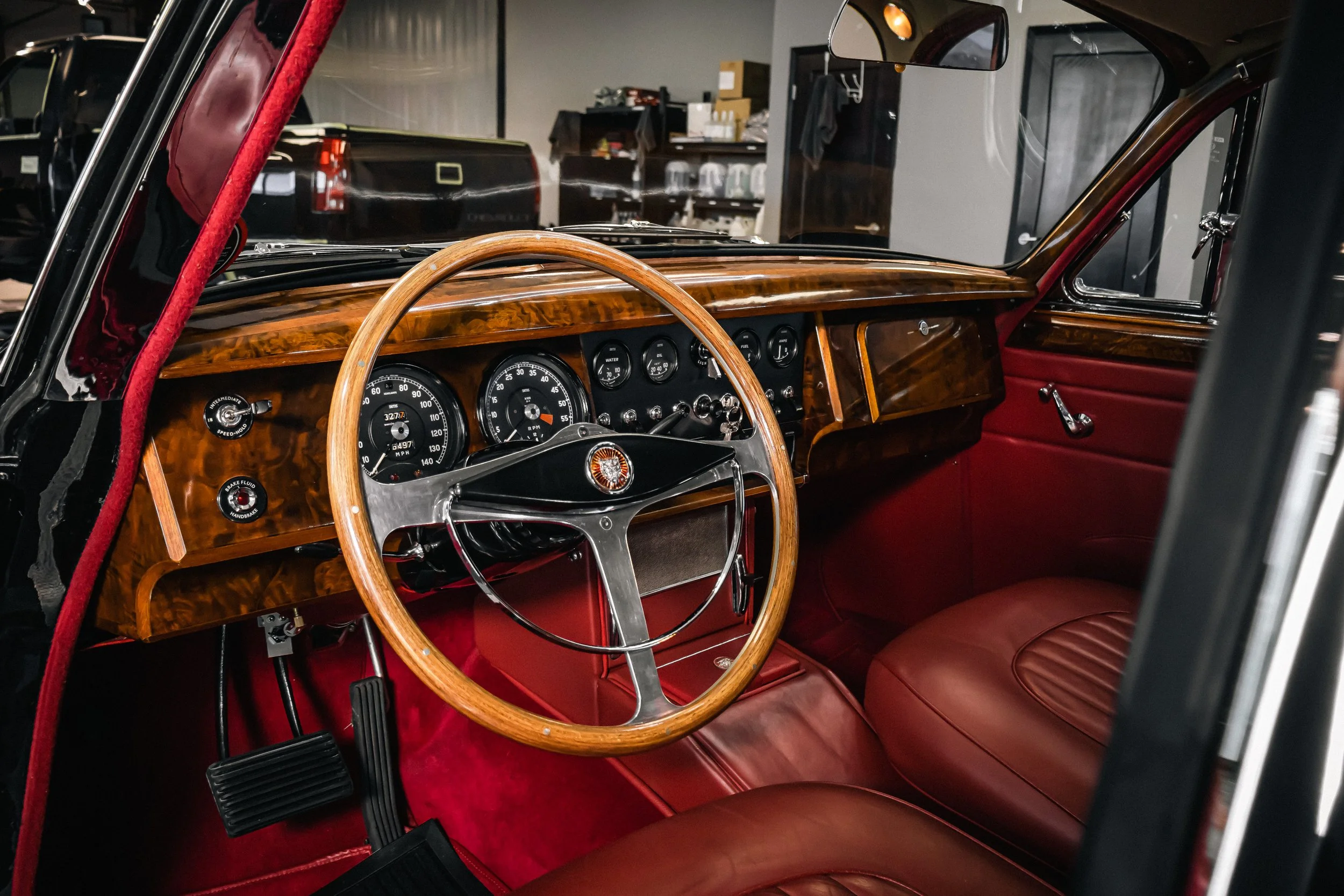 Interior of a vintage car showing a woodgrain dashboard, steering wheel, and red leather seats, with other vehicles and storage shelves visible in the background.