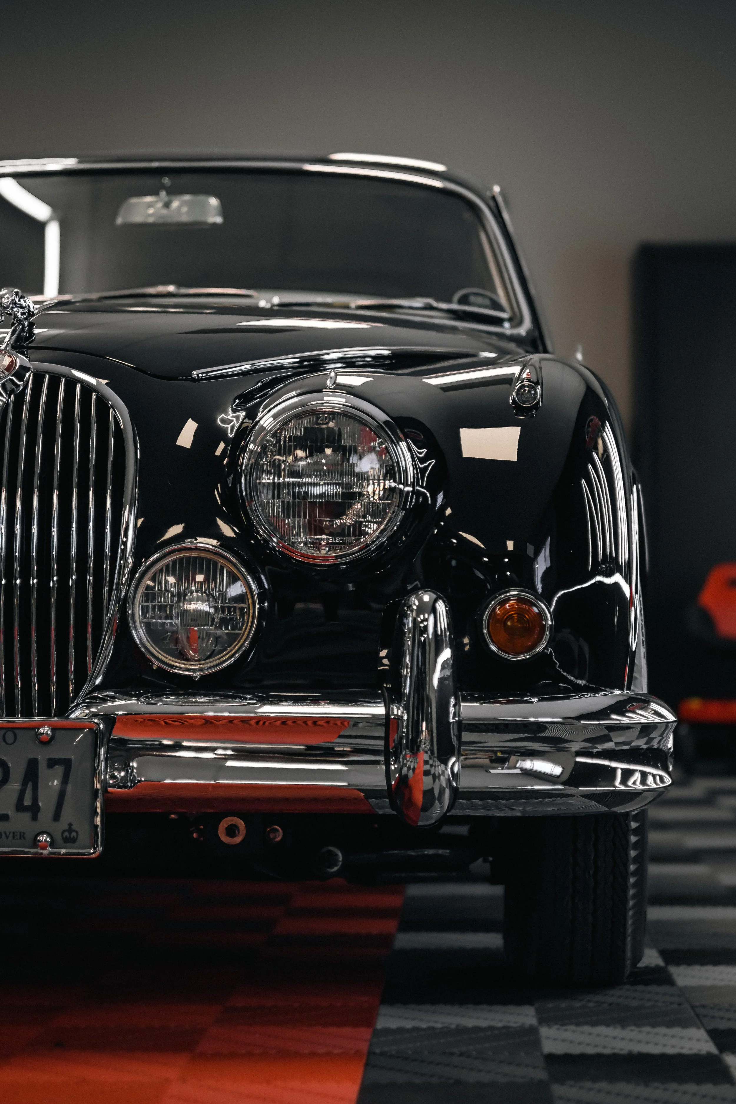Close-up of a vintage black car with chrome accents, featuring round headlights and chrome grill, parked on a checkered floor with red and black patterns.