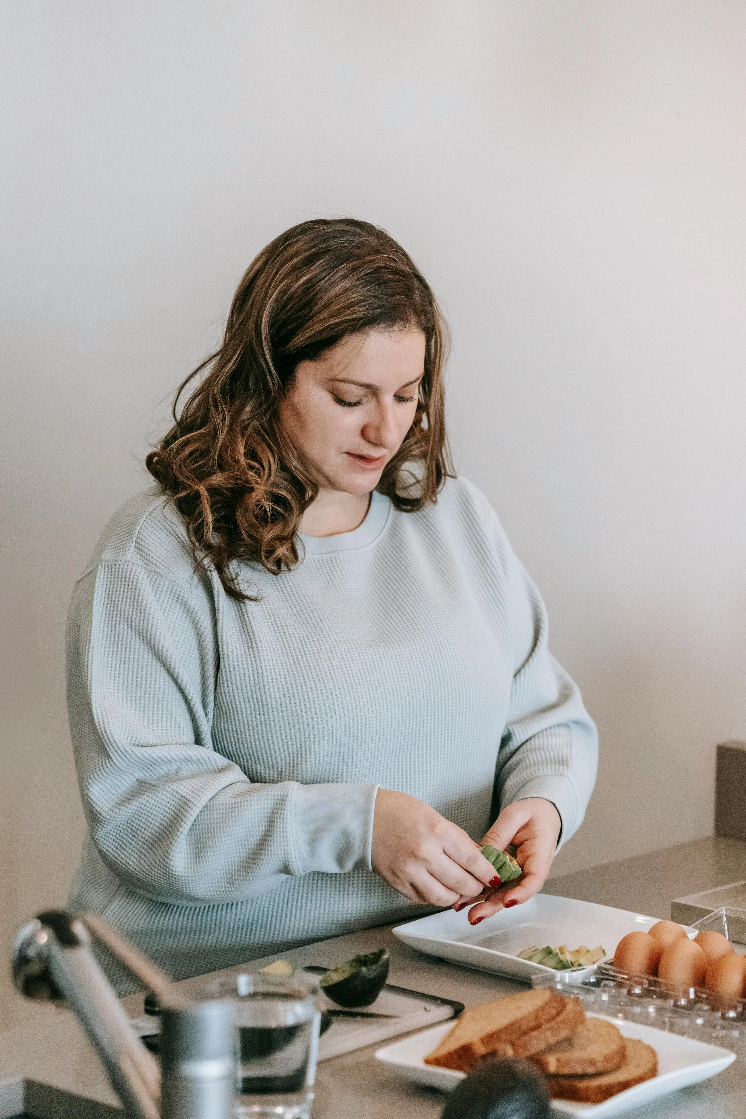 Woman preparing avocado slices on a kitchen counter, with eggs and bread nearby.