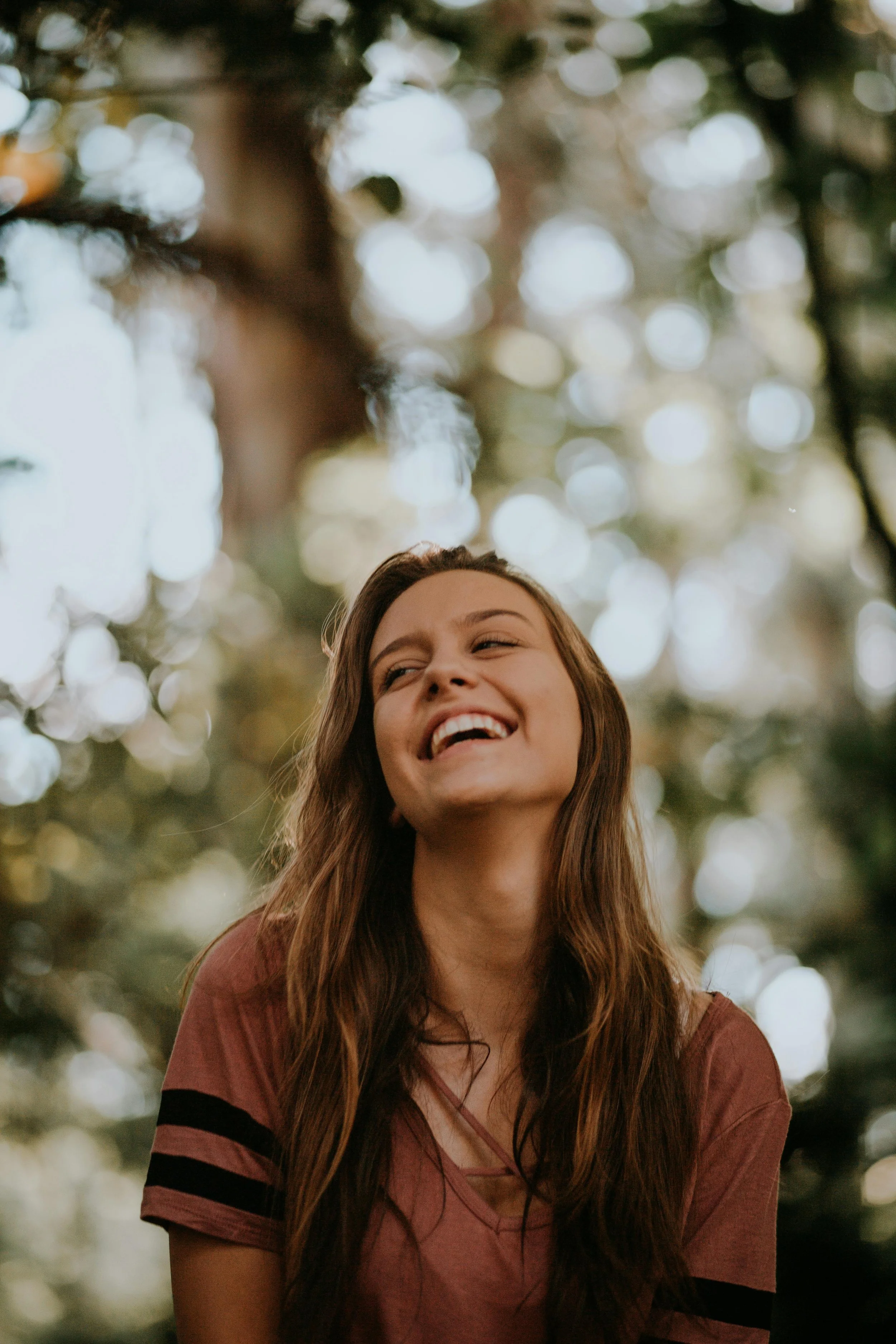 A young woman with long brown hair smiling and looking up outdoors in a forest with sunlight filtering through trees.