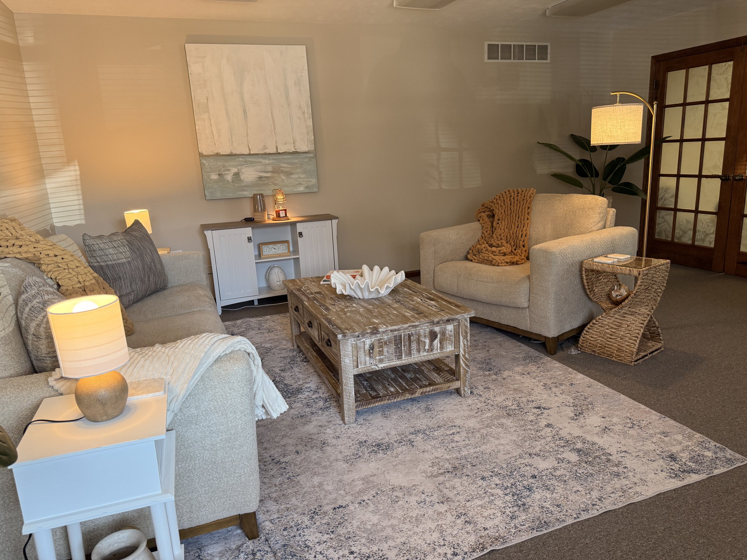 A cozy living room with beige sofas, a wooden coffee table, a white side table with a lamp, a woven chair, and a white cabinet against a gray wall with a large abstract painting, warm lighting, and a large plant in the corner.