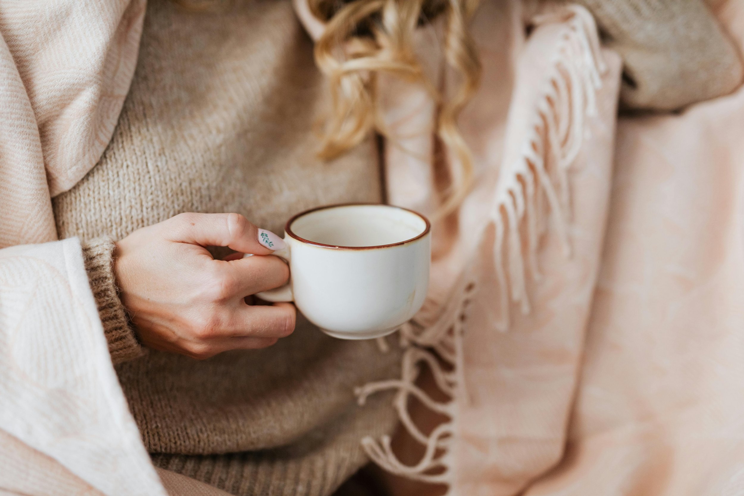 Person holding a white ceramic mug with brown trim while sitting on a beige sofa with a blanket.