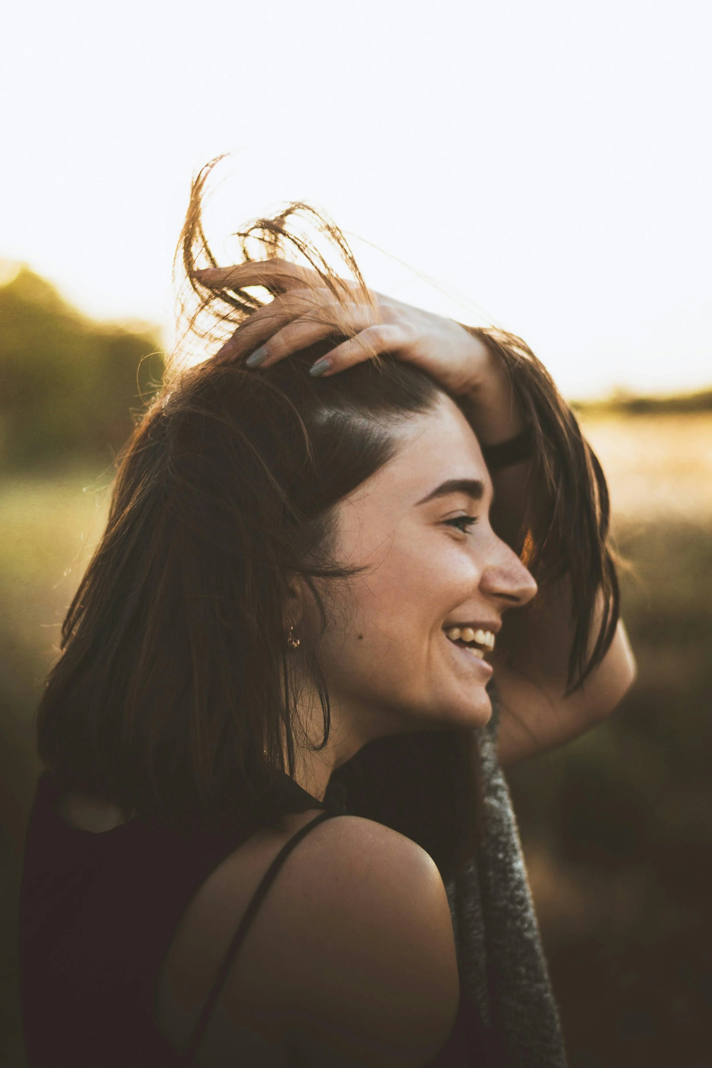 A young woman smiling outdoors with her eyes closed, holding her hair back with one hand, during sunset.