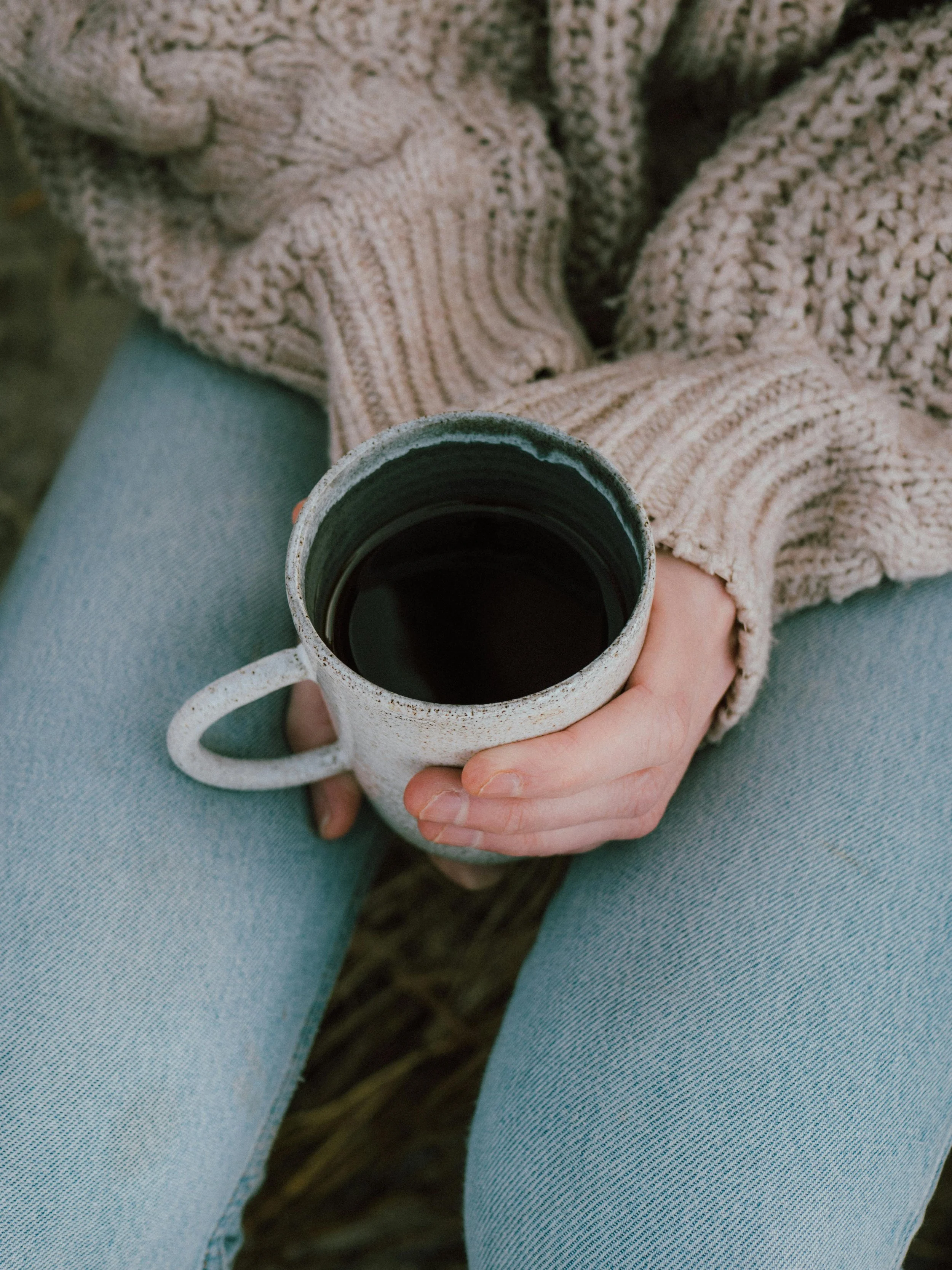 Person sitting outdoors holding a ceramic mug filled with a dark beverage, wearing light blue jeans and a chunky beige knit sweater.