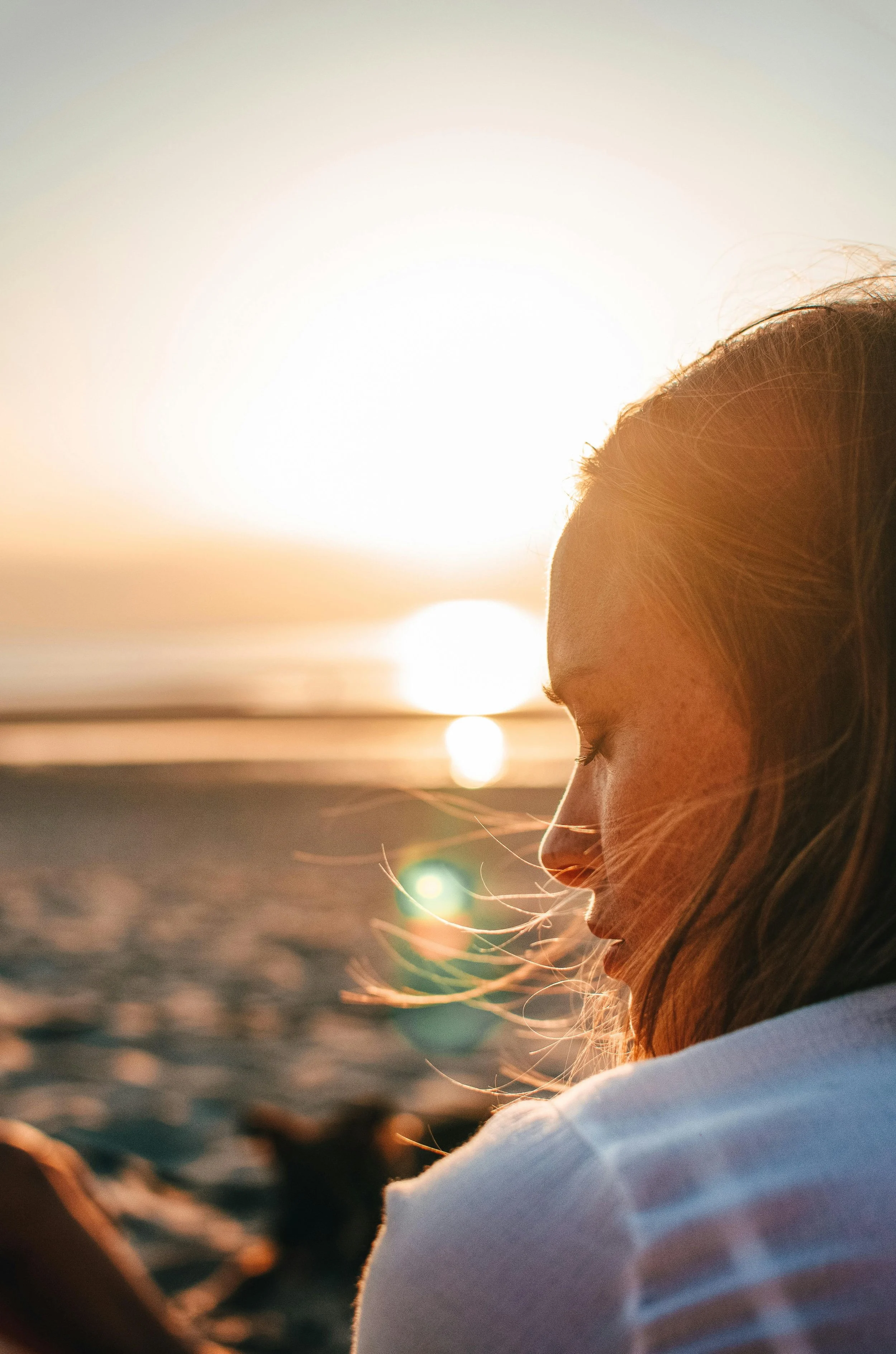 A woman with reddish hair and freckles sits on a beach at sunset, with her eyes closed and head slightly bowed, the sun low in the sky behind her.