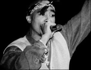 Tupac Shakur wearing a bandana and a denim vest, holding a microphone to his mouth with one arm raised in a dynamic performance pose against a dark background.