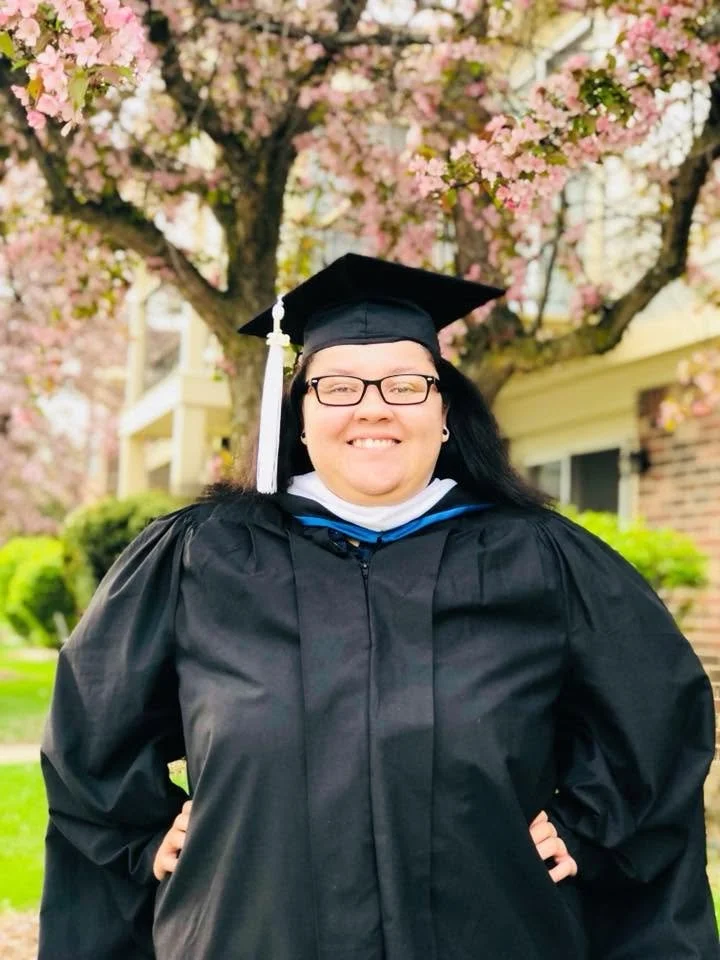 Holly E. Burgess in graduation robes and cap smiling outdoors with pink cherry blossoms and a house in the background.