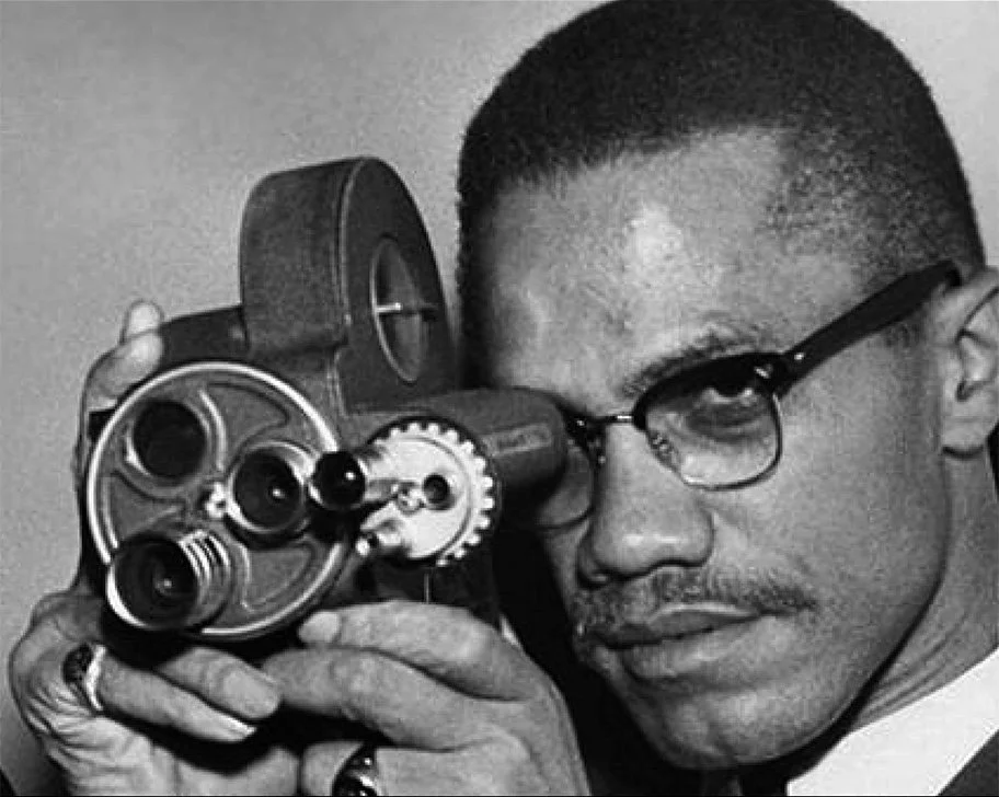 Close-up of Malcolm X wearing glasses and a suit, holding a professional 8mm film camera to his eye.