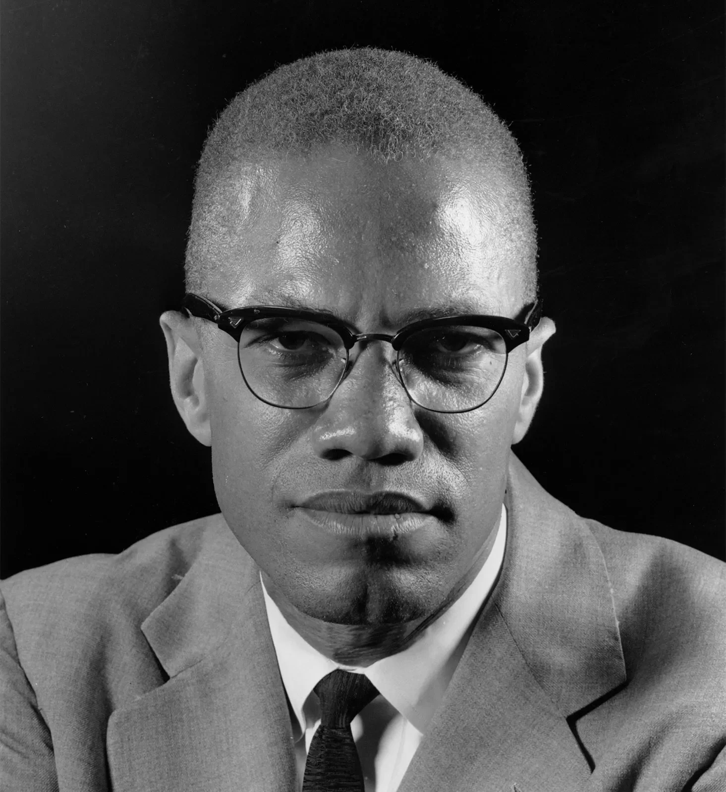 High-resolution portrait of Malcolm X wearing his signature horn-rimmed glasses, a suit jacket, and a dark tie against a black background.